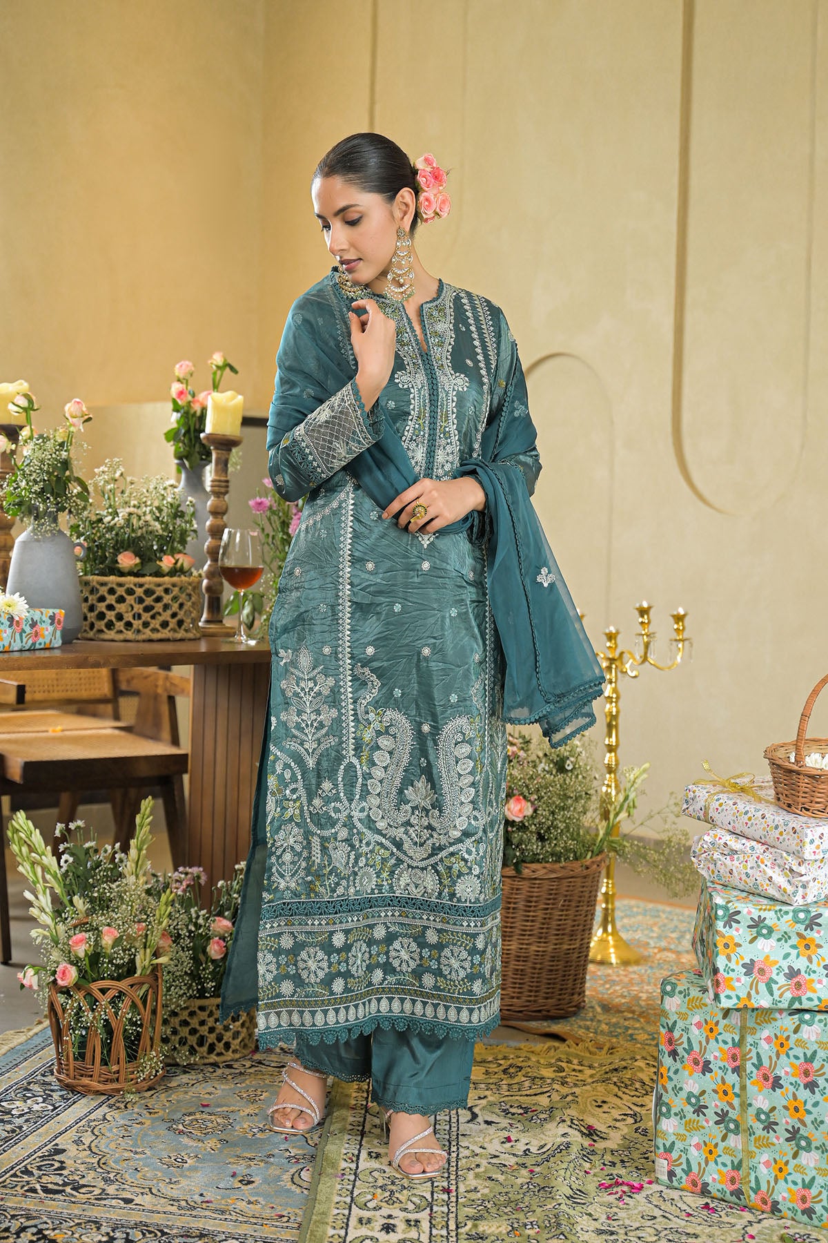 Woman in teal embroidered outfit standing in a decorated room with flowers and furniture.