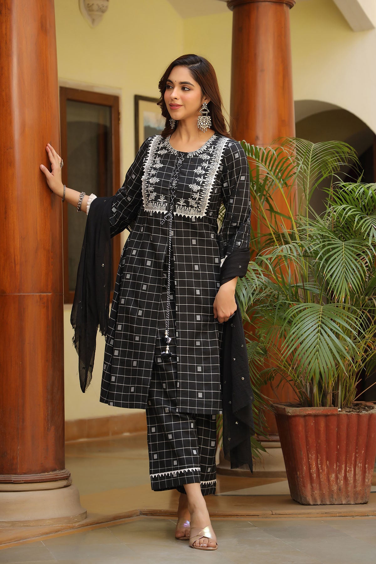 Woman in a black and white checkered outfit standing indoors with plants and wooden columns in the background.