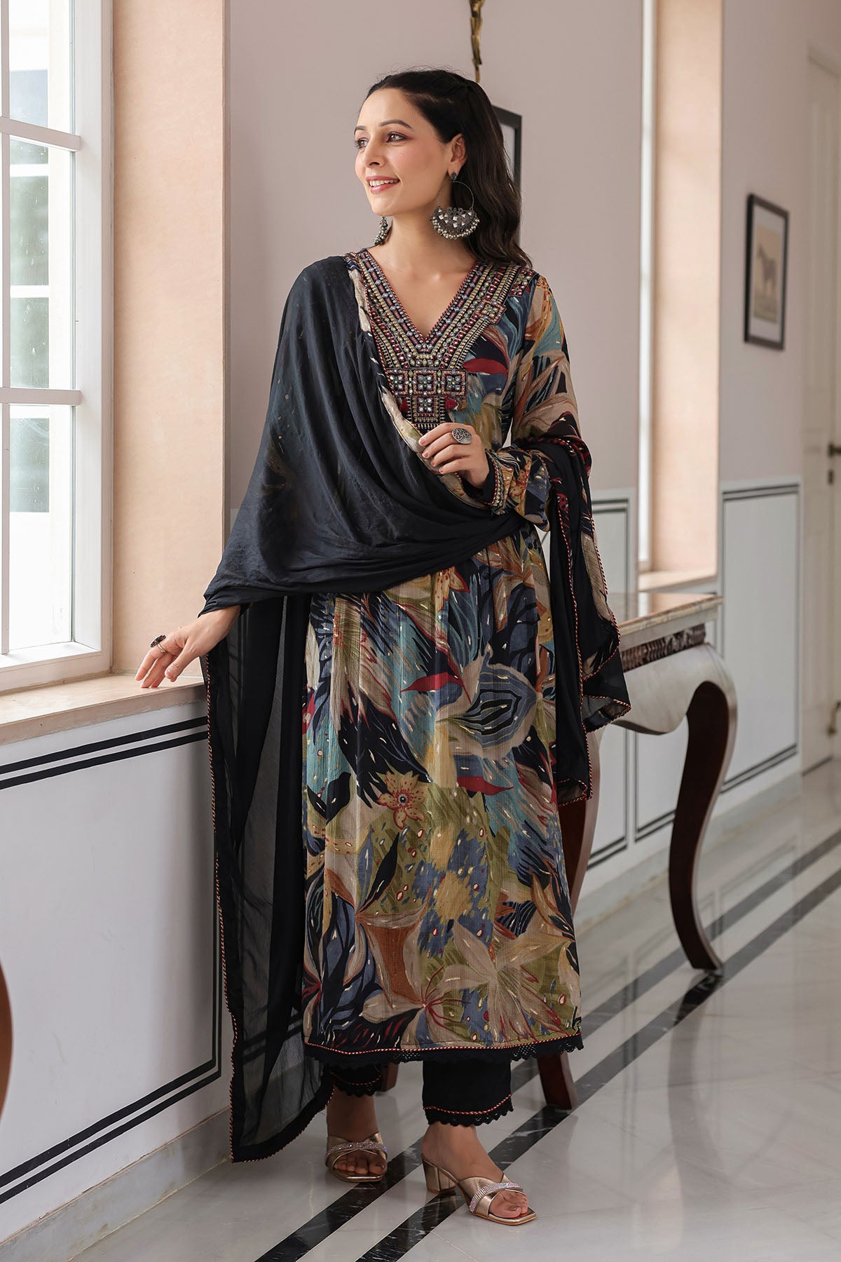 Woman in a colorful traditional outfit standing in a room with a window and chair.