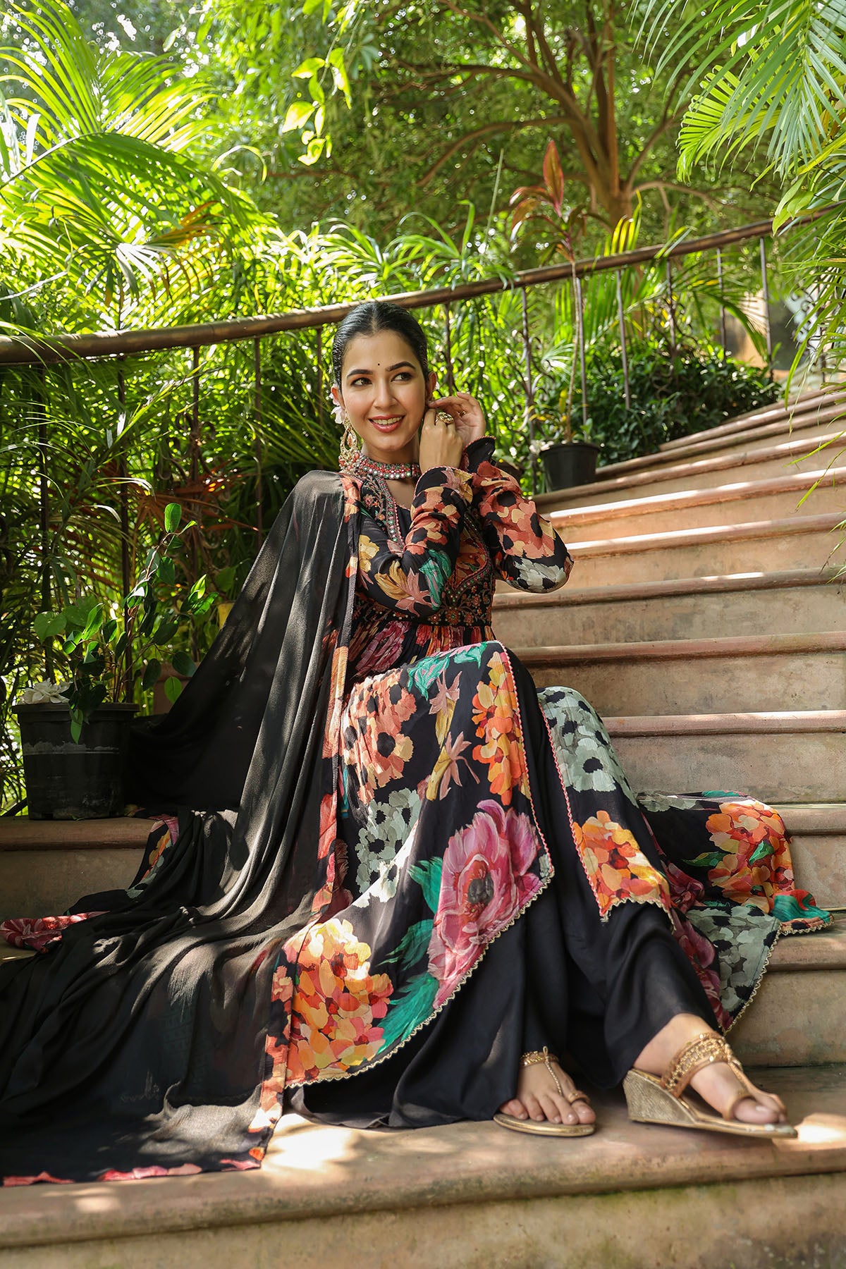 Woman in a floral and black traditional outfit sitting on steps with greenery in the background