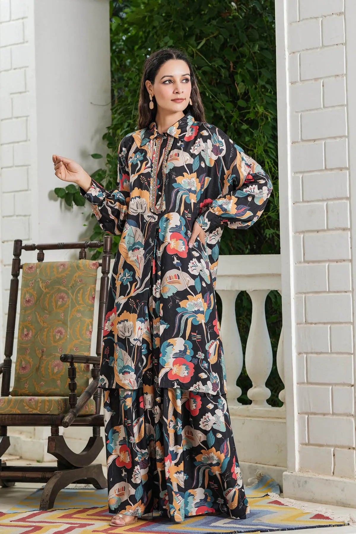 Woman wearing a floral dress standing on a porch with a chair and plants in the background.
