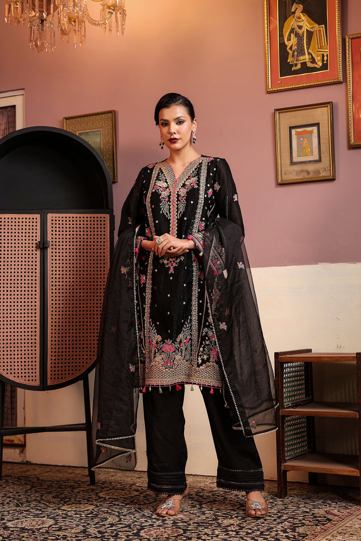 Woman in traditional black and gold embroidered outfit standing in a decorated room with paintings on the wall.