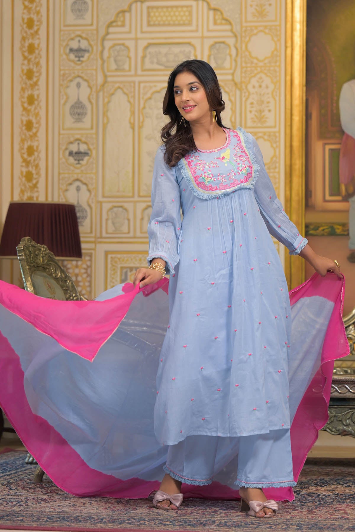 Woman in a light blue traditional outfit with pink dupatta standing in a decorated room.