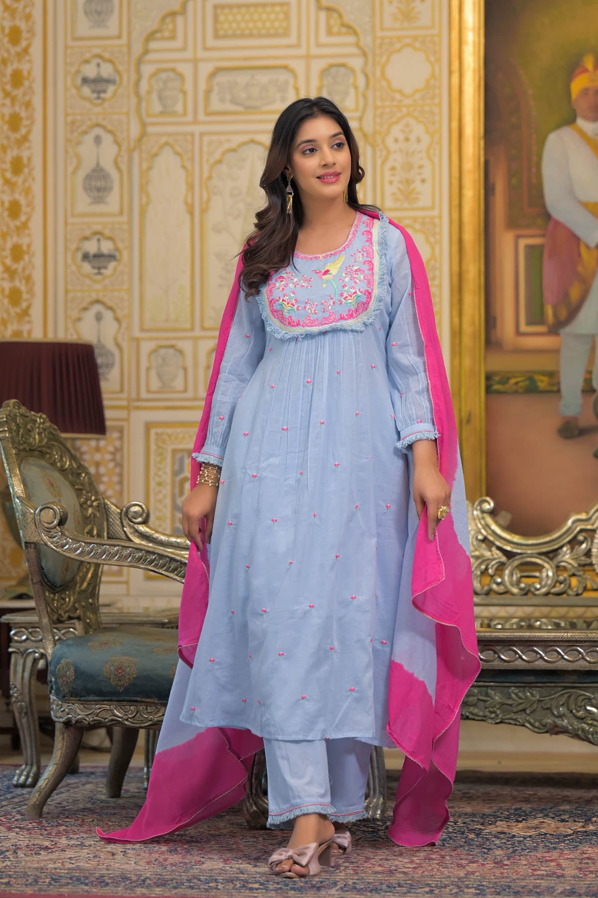 Woman in traditional outfit with pink dupatta standing in a decorated room.