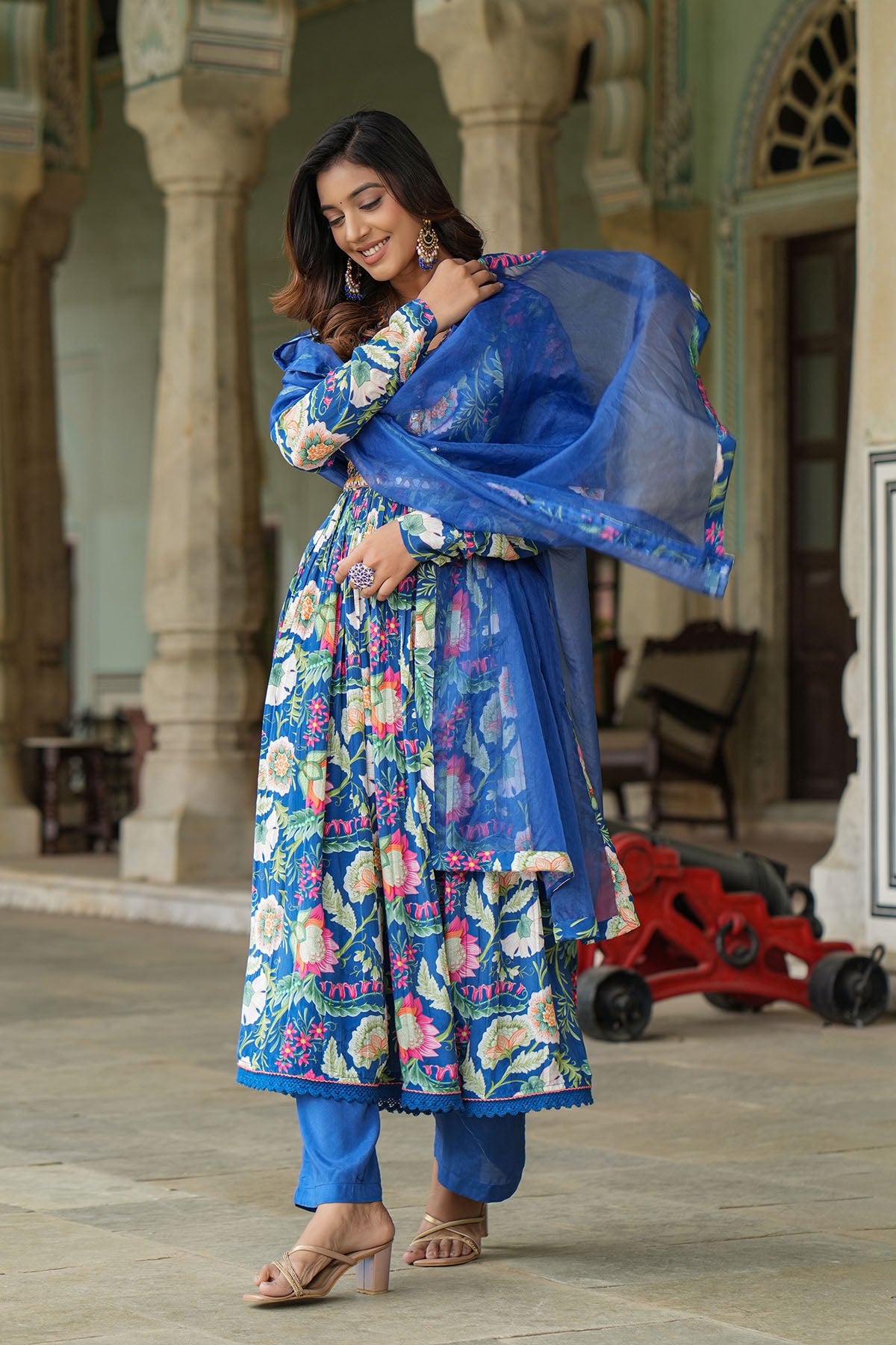 Woman in a blue floral traditional outfit standing in an outdoor setting with architectural columns.