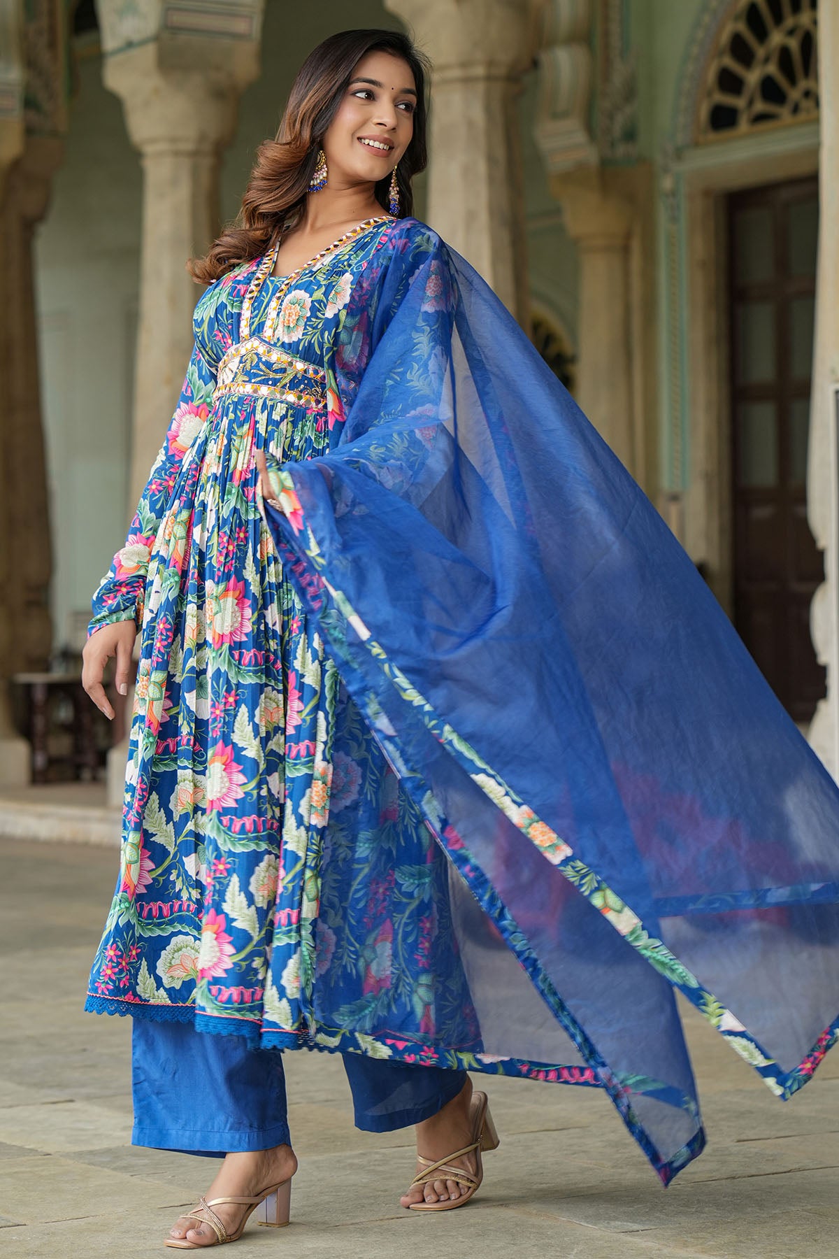 Woman wearing a colorful floral dress with a blue dupatta in an outdoor setting.