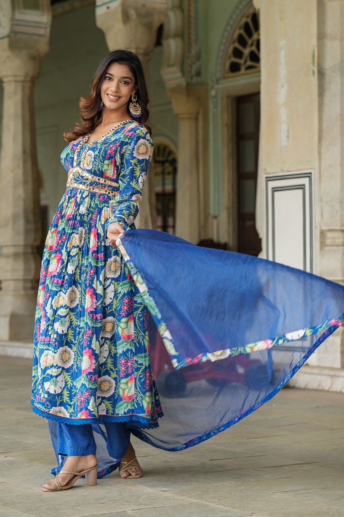 Woman in a colorful floral dress with a blue dupatta standing in front of an architectural background.