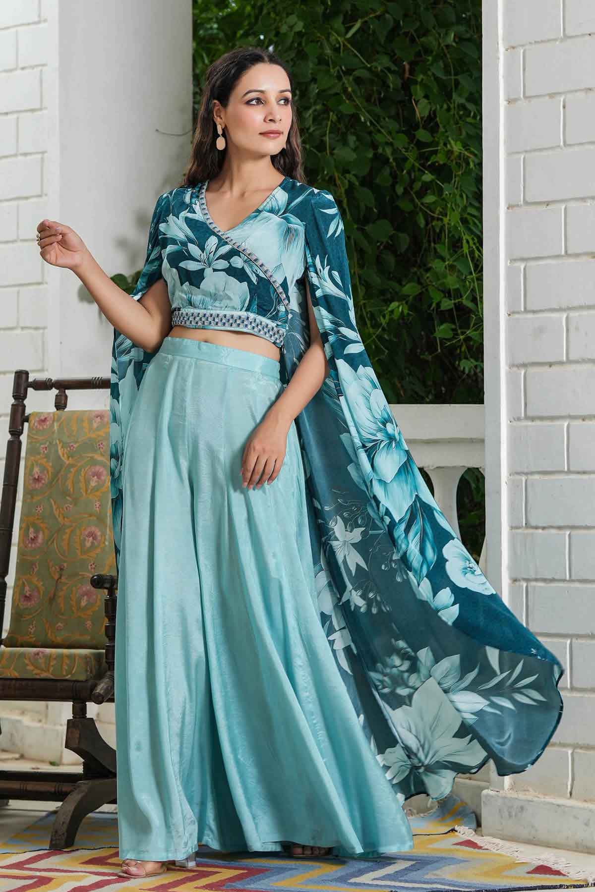 Woman in a teal floral saree standing outdoors with a white column and greenery in the background.