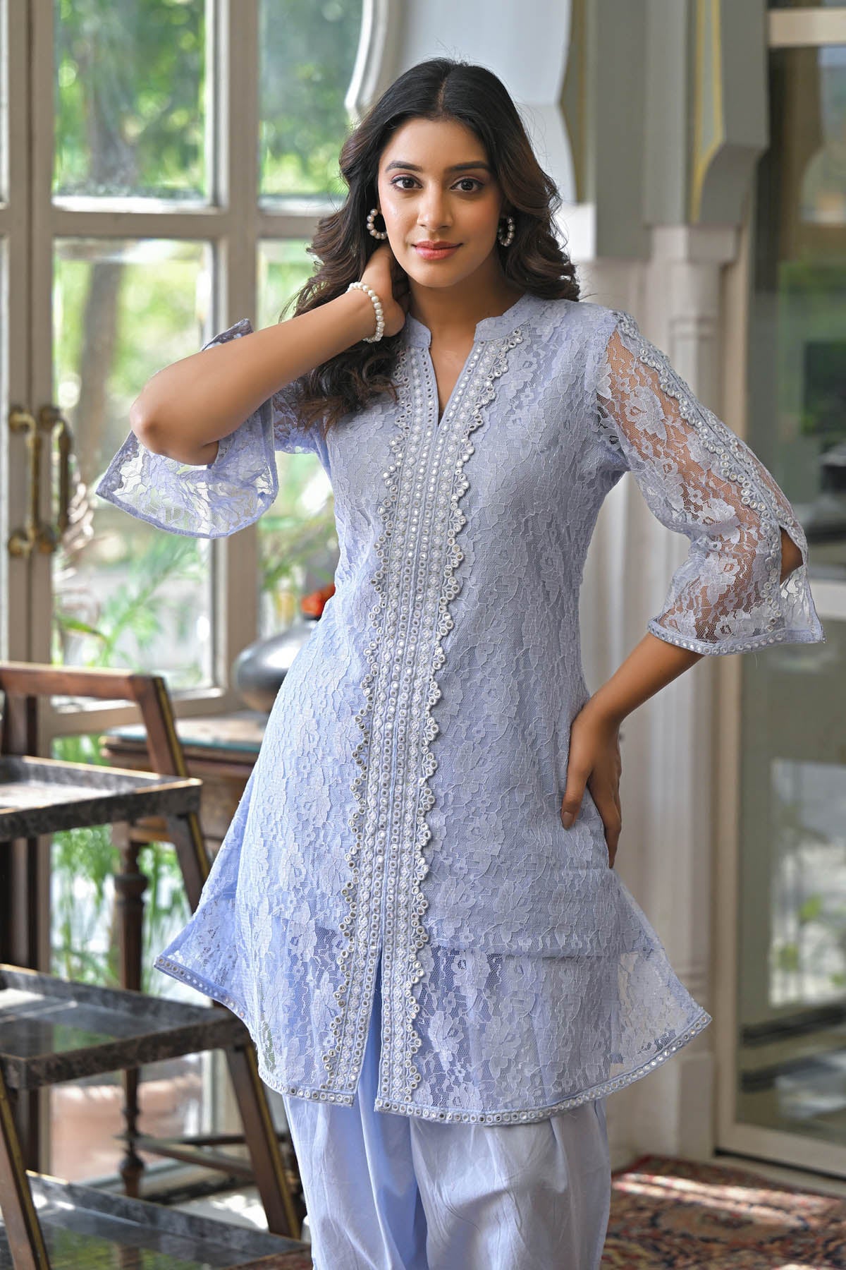 Woman wearing a light blue embroidered outfit in a room with large windows.