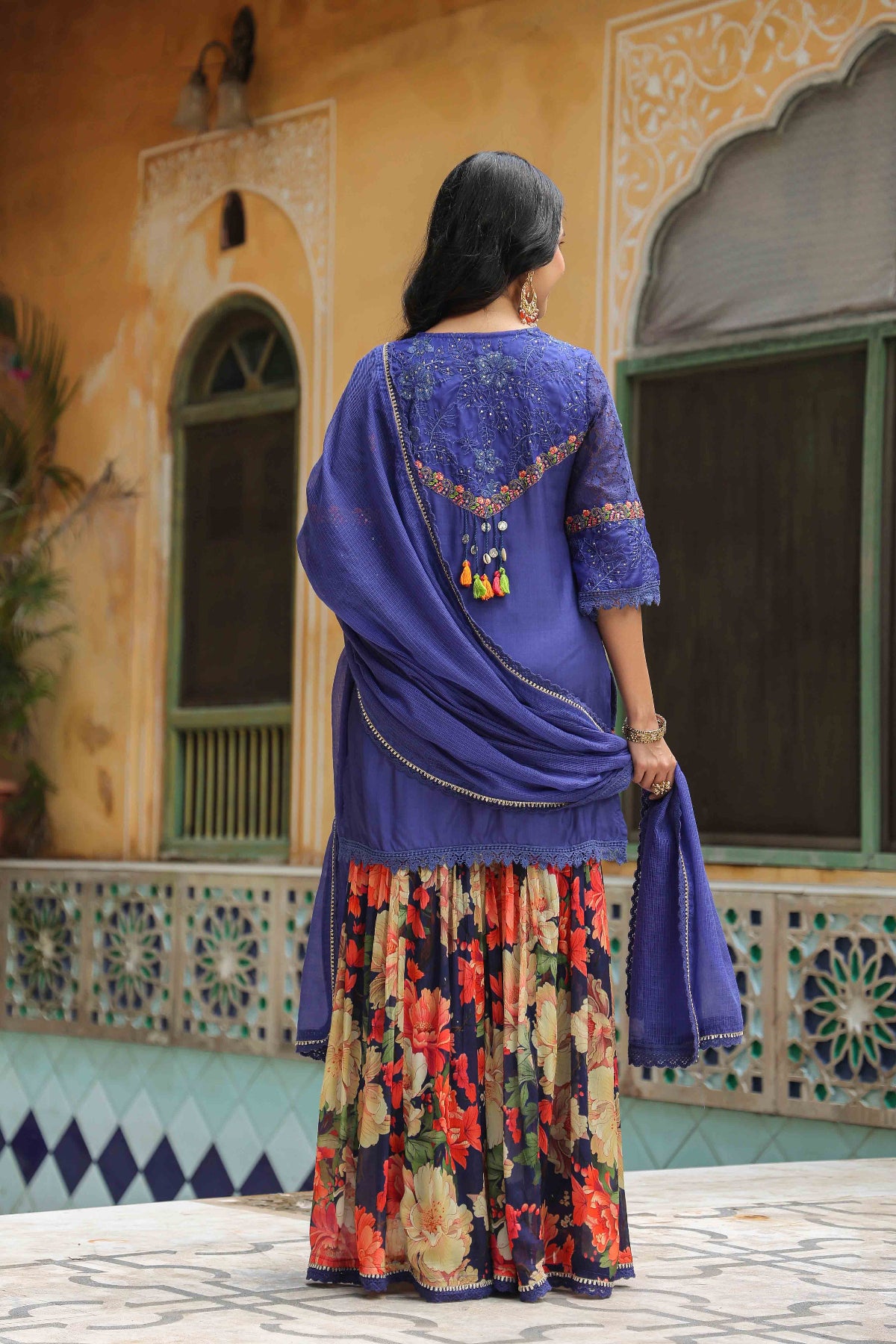 Woman in a blue embroidered top and colorful skirt standing in front of a decorative building.