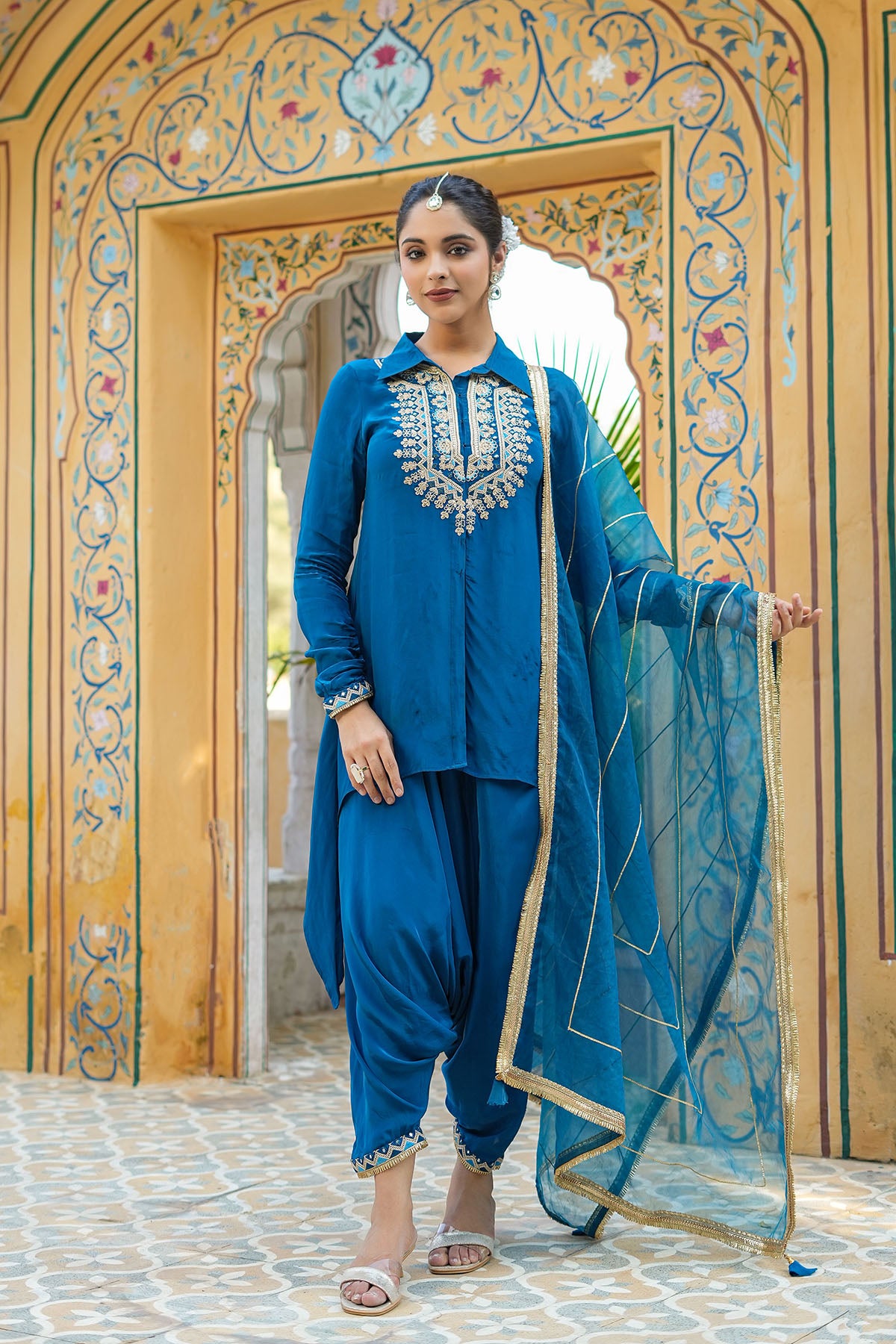 Woman in a blue traditional outfit standing in front of an ornate wooden door.