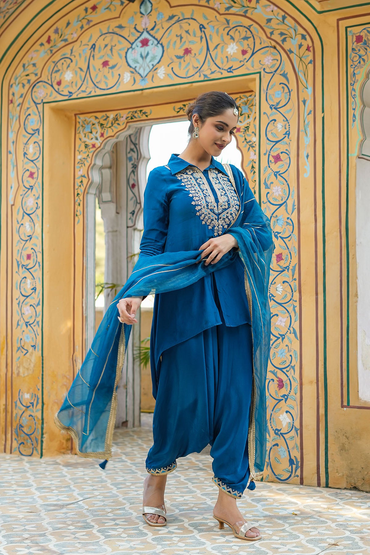 Woman in a blue traditional outfit standing in front of an ornate archway.