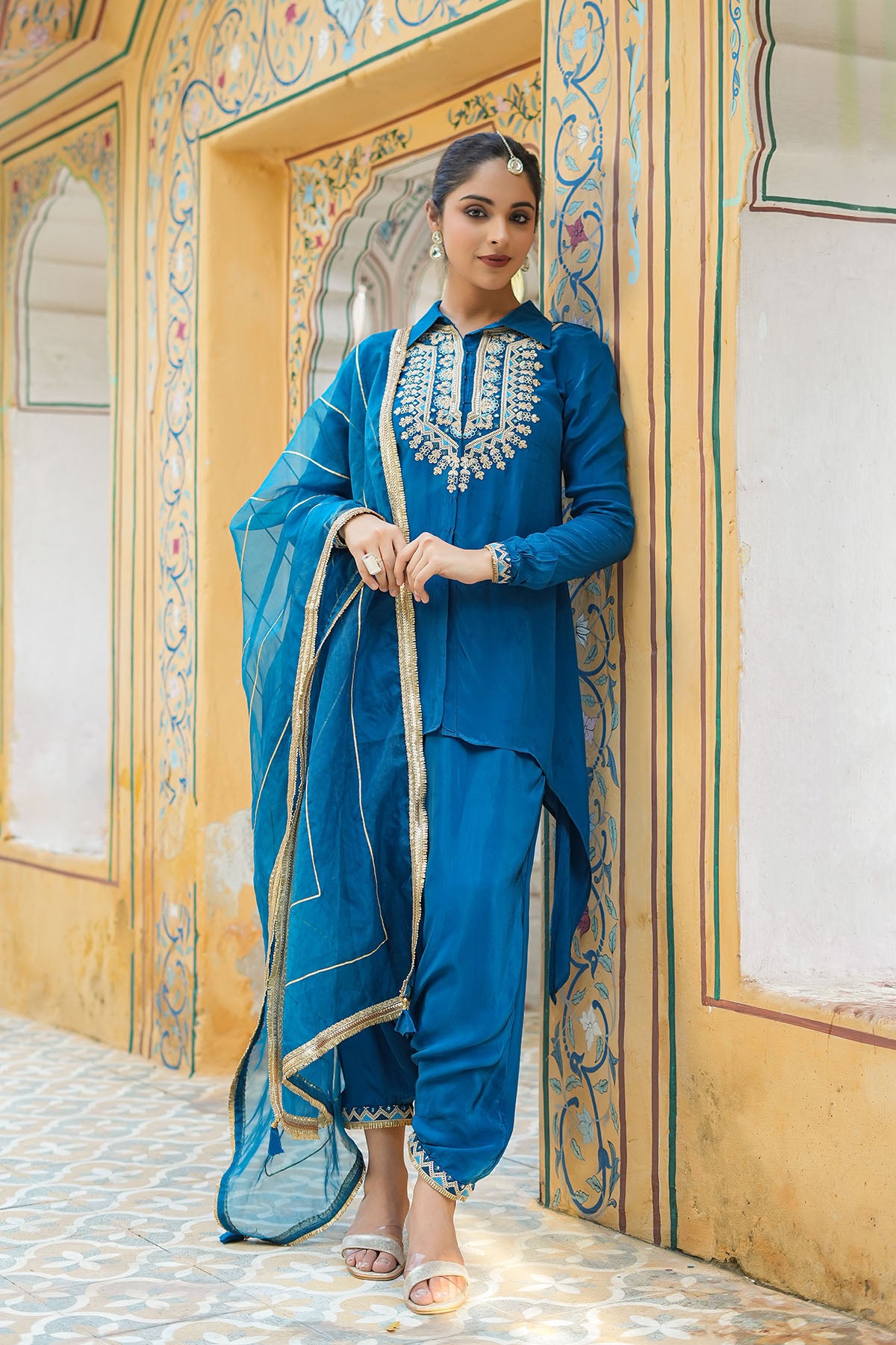 Woman in a blue traditional outfit standing in front of an ornate wooden door.