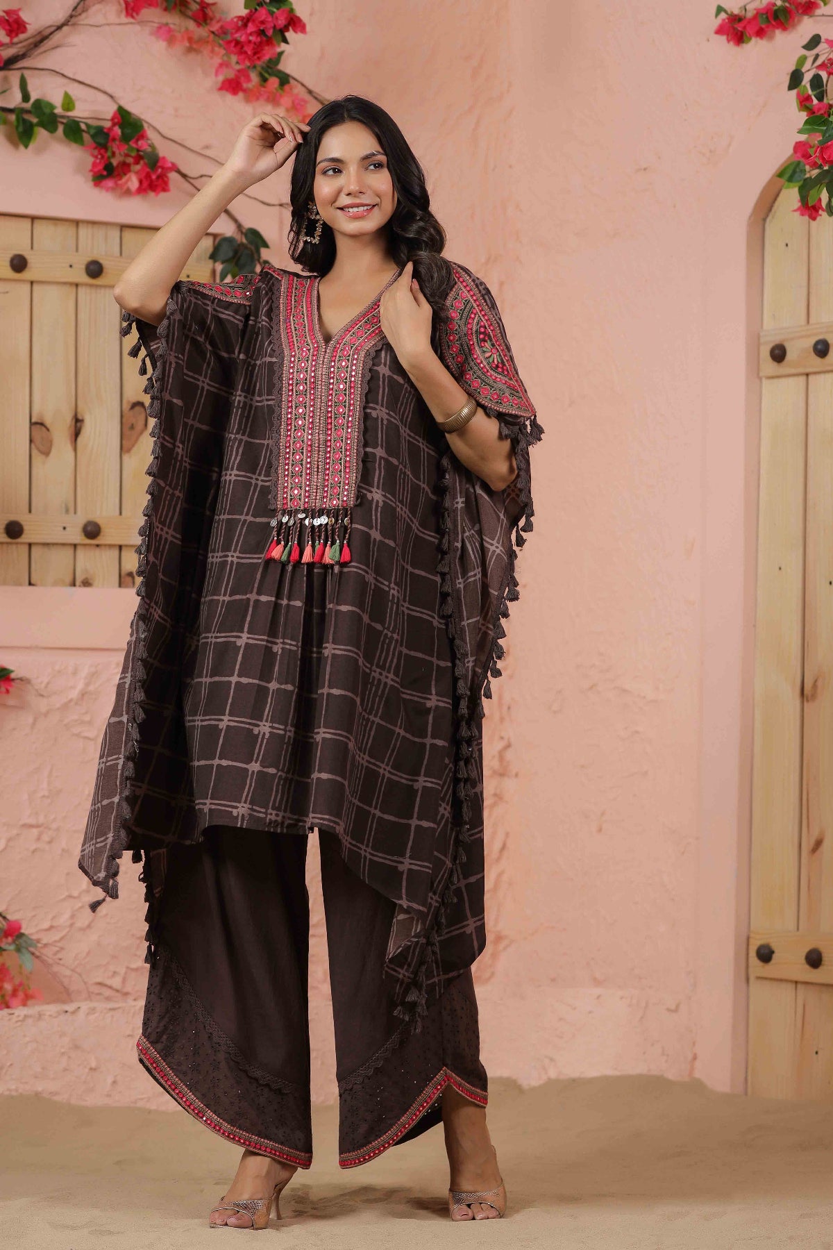 Woman wearing a traditional outfit with a checkered shawl and black pants against a pink wall.