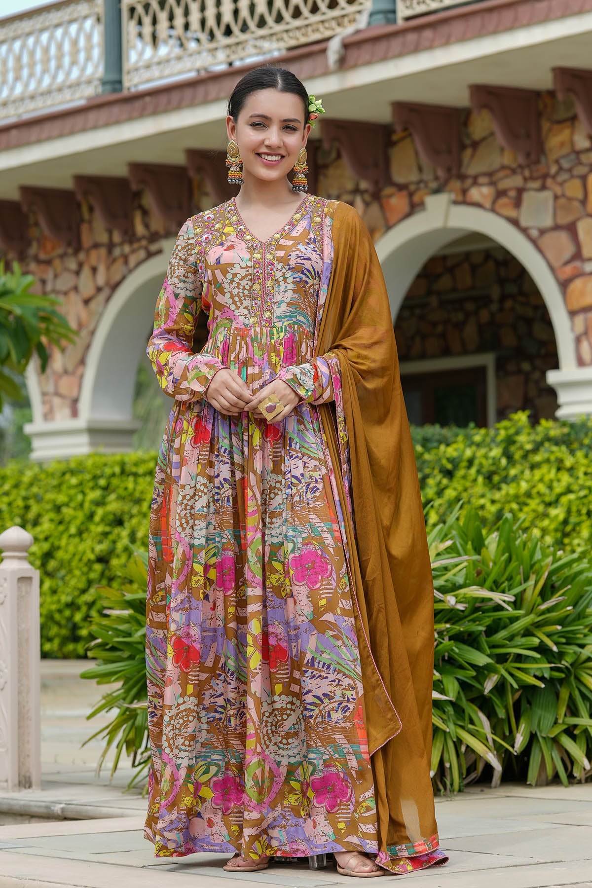 Woman in a colorful traditional outfit standing outdoors with a stone building and greenery in the background