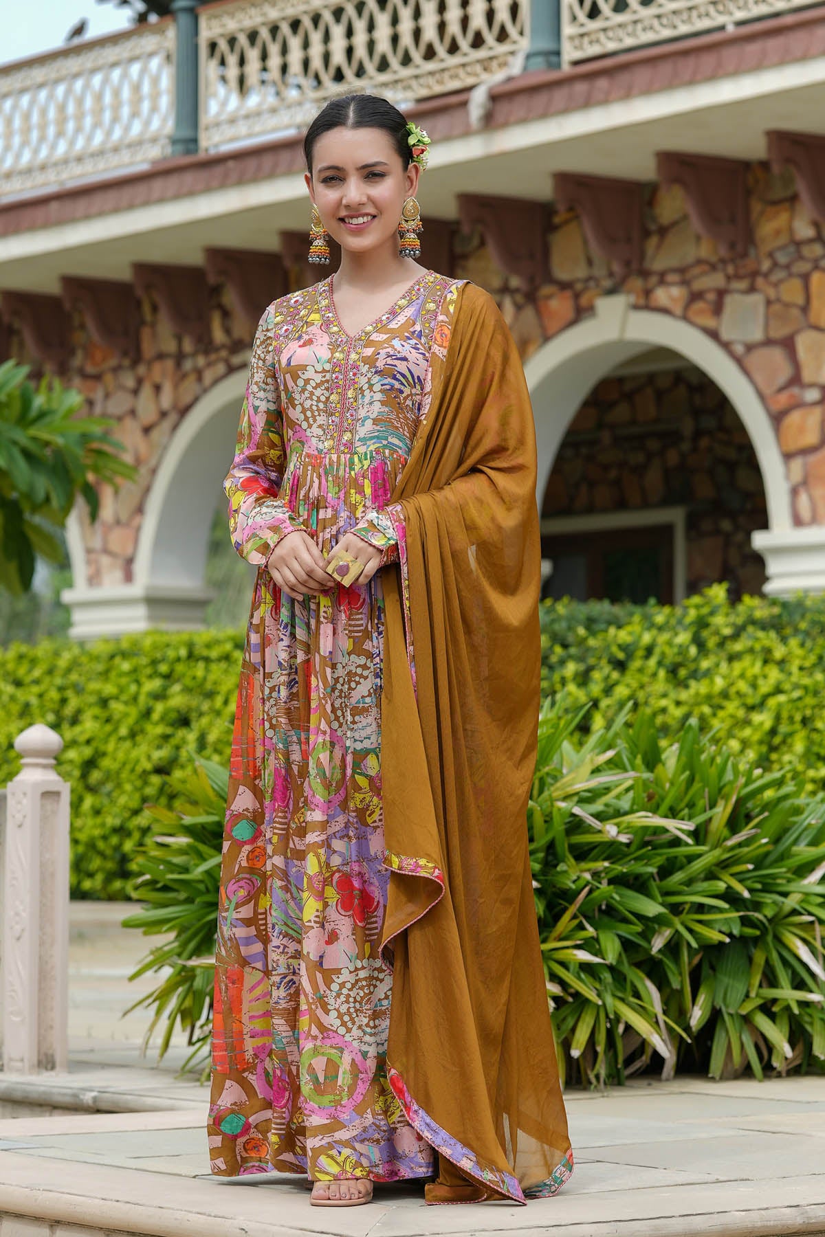Woman in a colorful traditional outfit standing outdoors with greenery and a building in the background