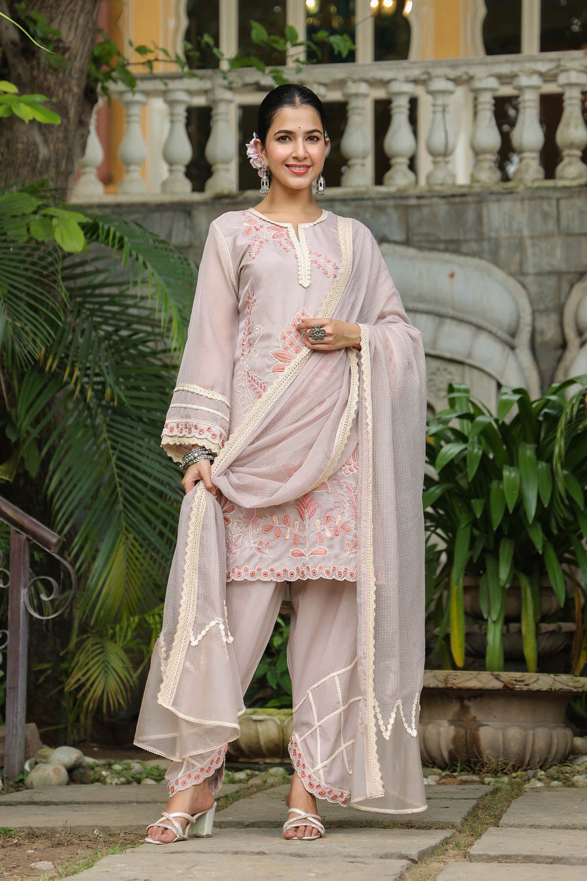Woman in a traditional outfit standing outdoors with greenery and architectural elements in the background