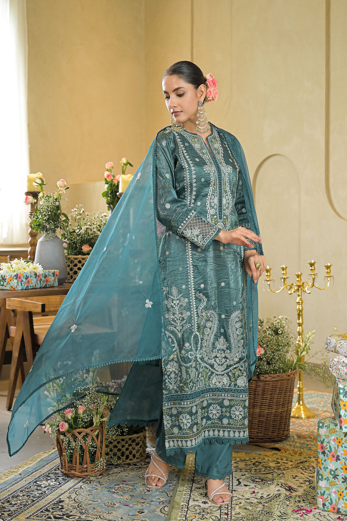 Woman in a teal embroidered outfit standing in an elegant room with floral arrangements and a candelabra.