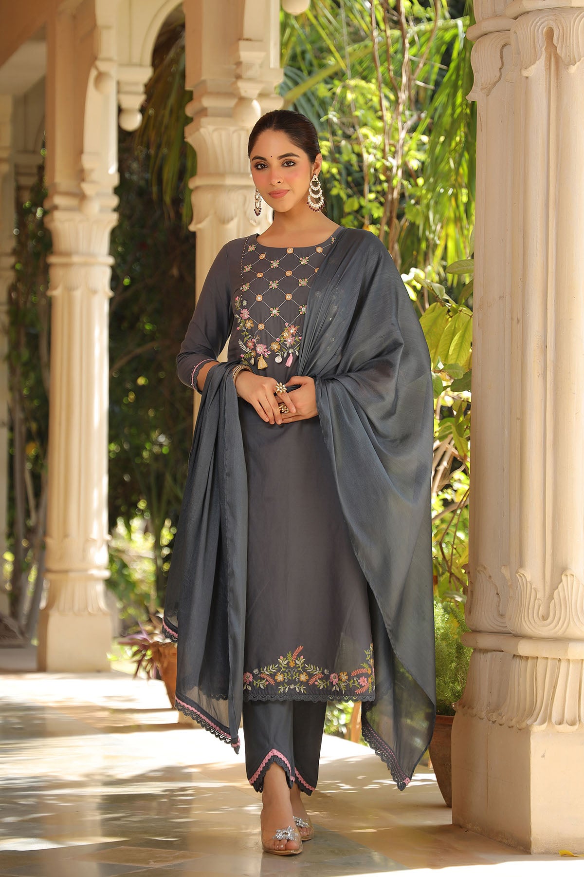 Woman in a dark gray traditional outfit with a floral shawl standing in an outdoor setting.