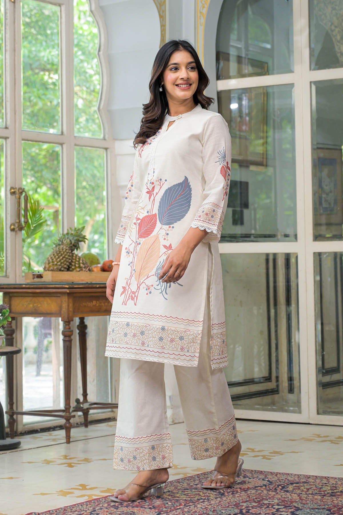 Woman in a white embroidered outfit standing in a room with large windows.