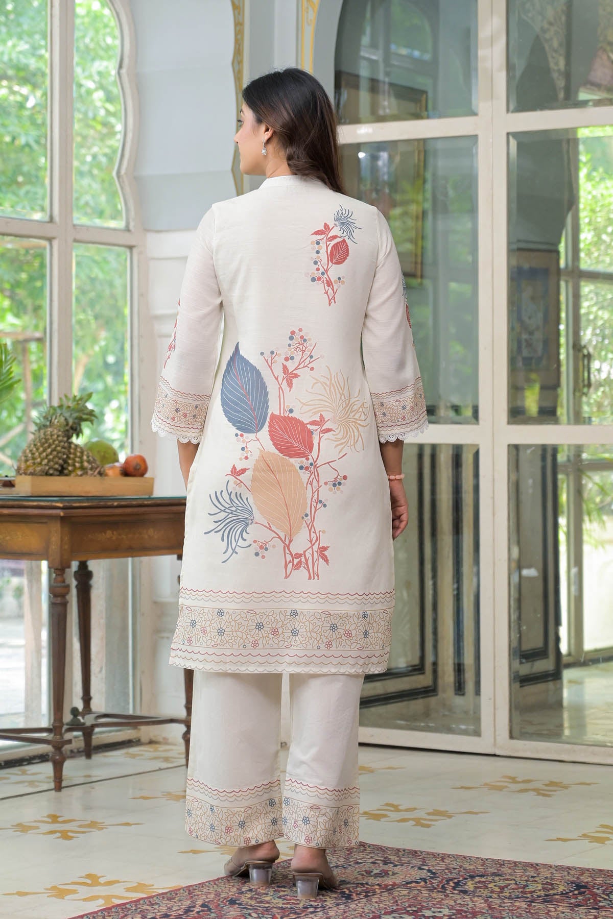Woman wearing a light-colored embroidered outfit in a room with a table and decorative items.