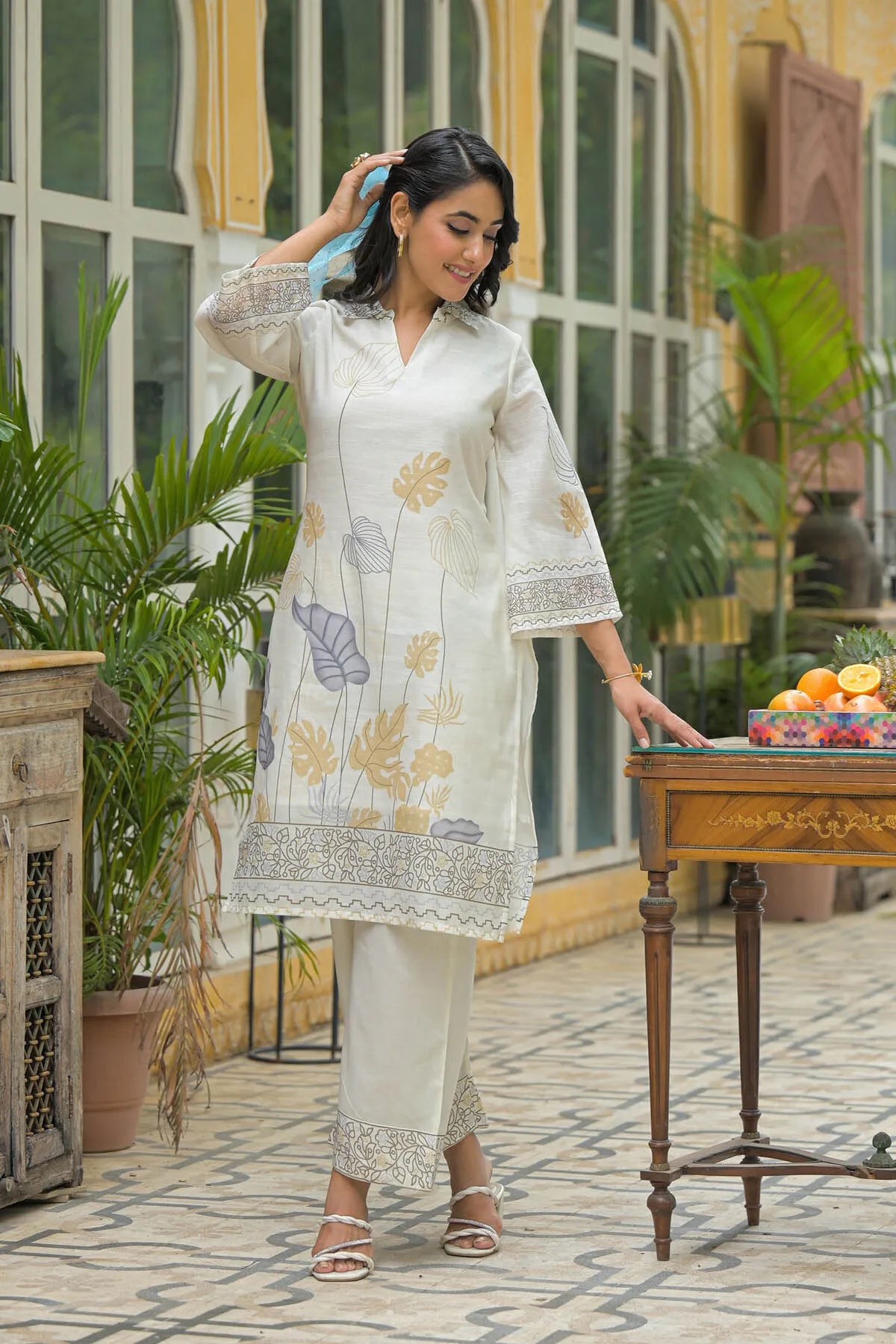 Woman in a white embroidered outfit standing in an outdoor setting with plants and a table.