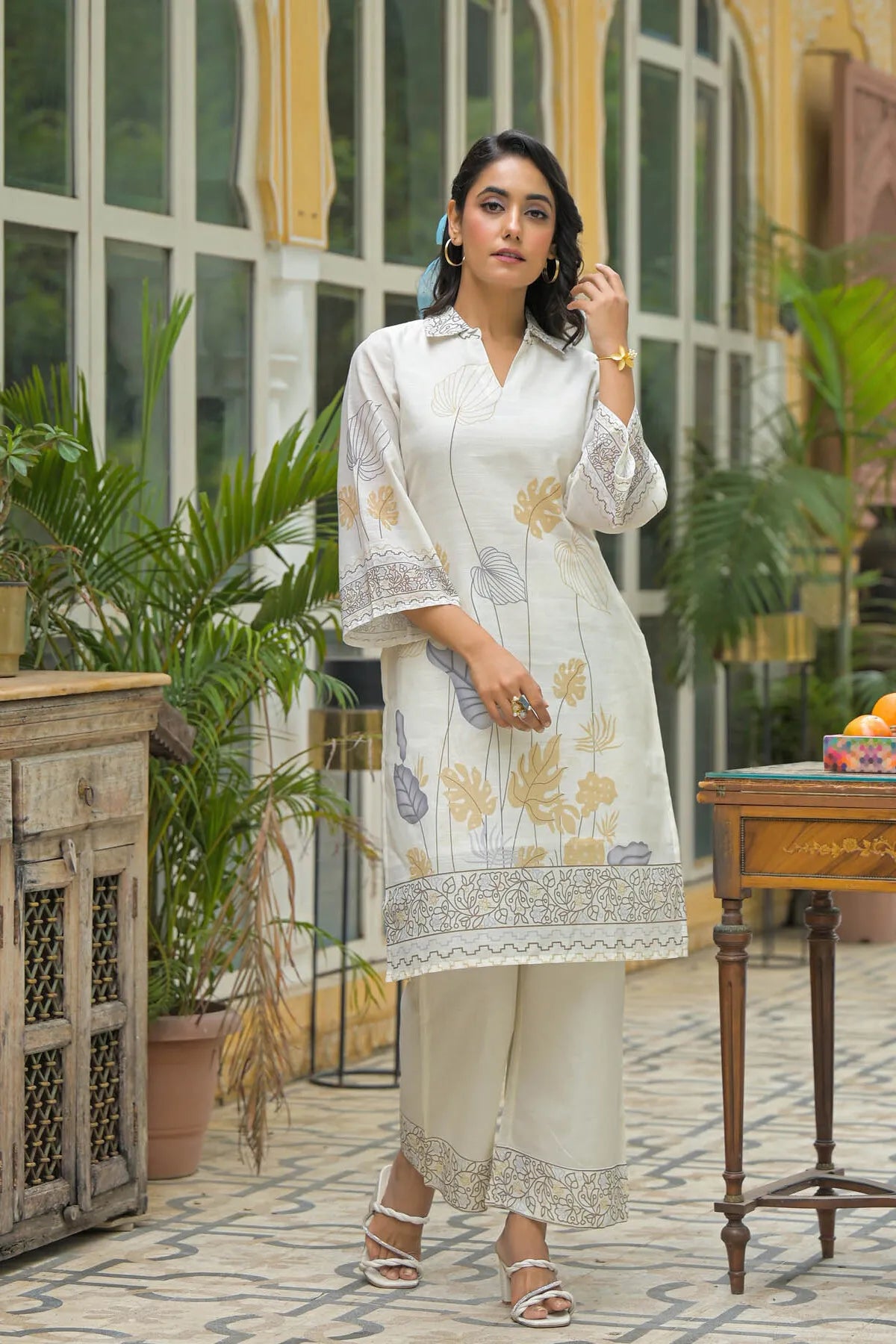 Woman in a white embroidered outfit standing in an outdoor setting with plants and furniture.