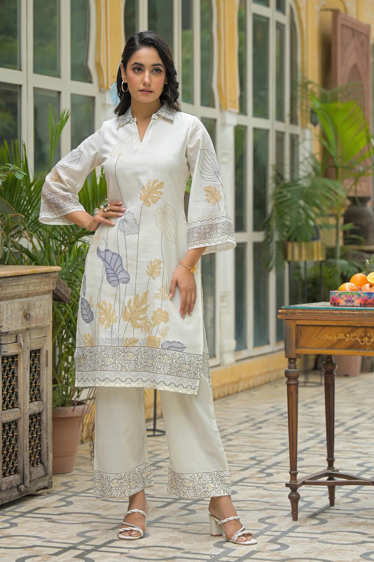 Woman in a white embroidered outfit standing in an outdoor setting with plants and a table.
