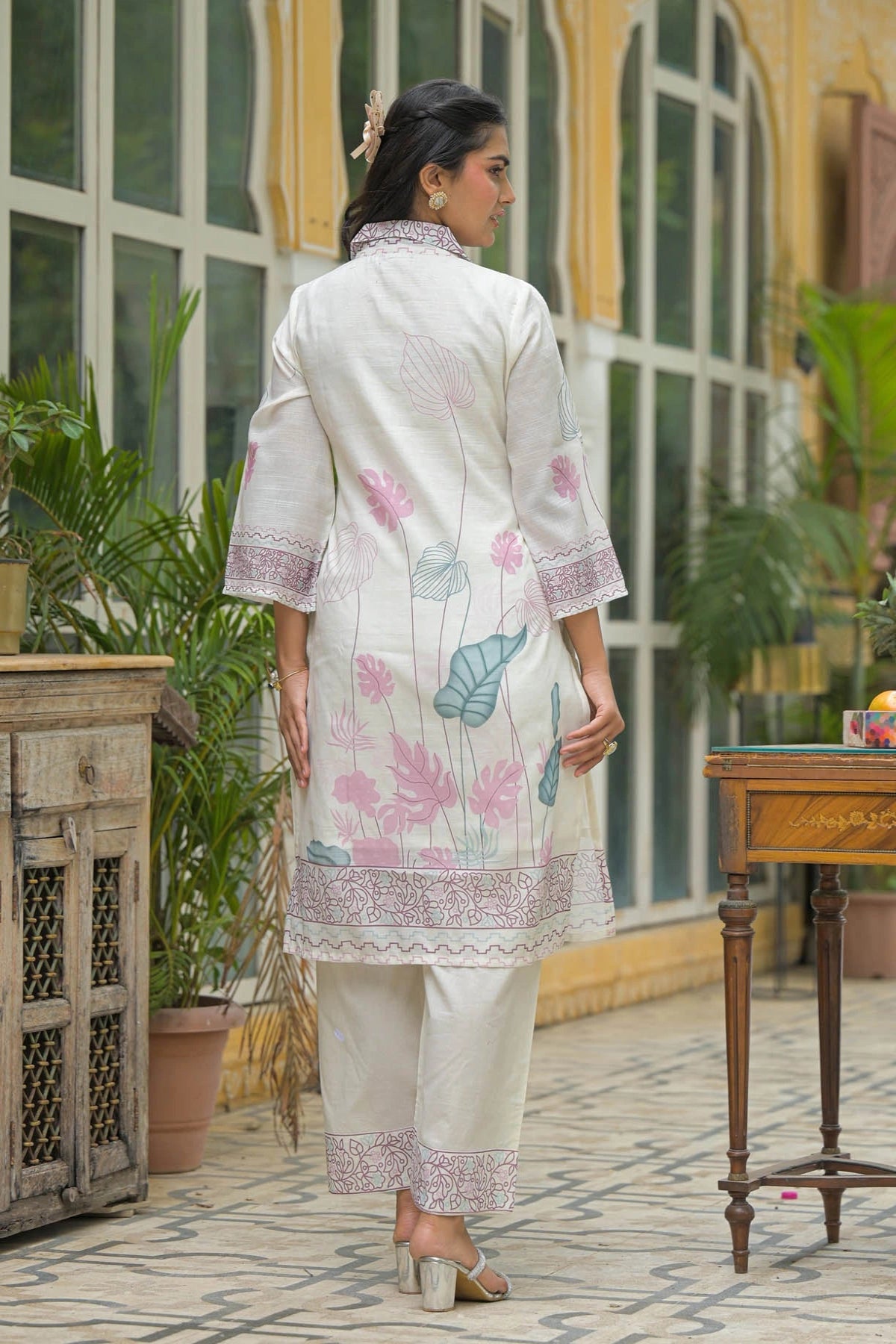Woman in a white floral outfit standing in a decorative indoor setting with plants and furniture.