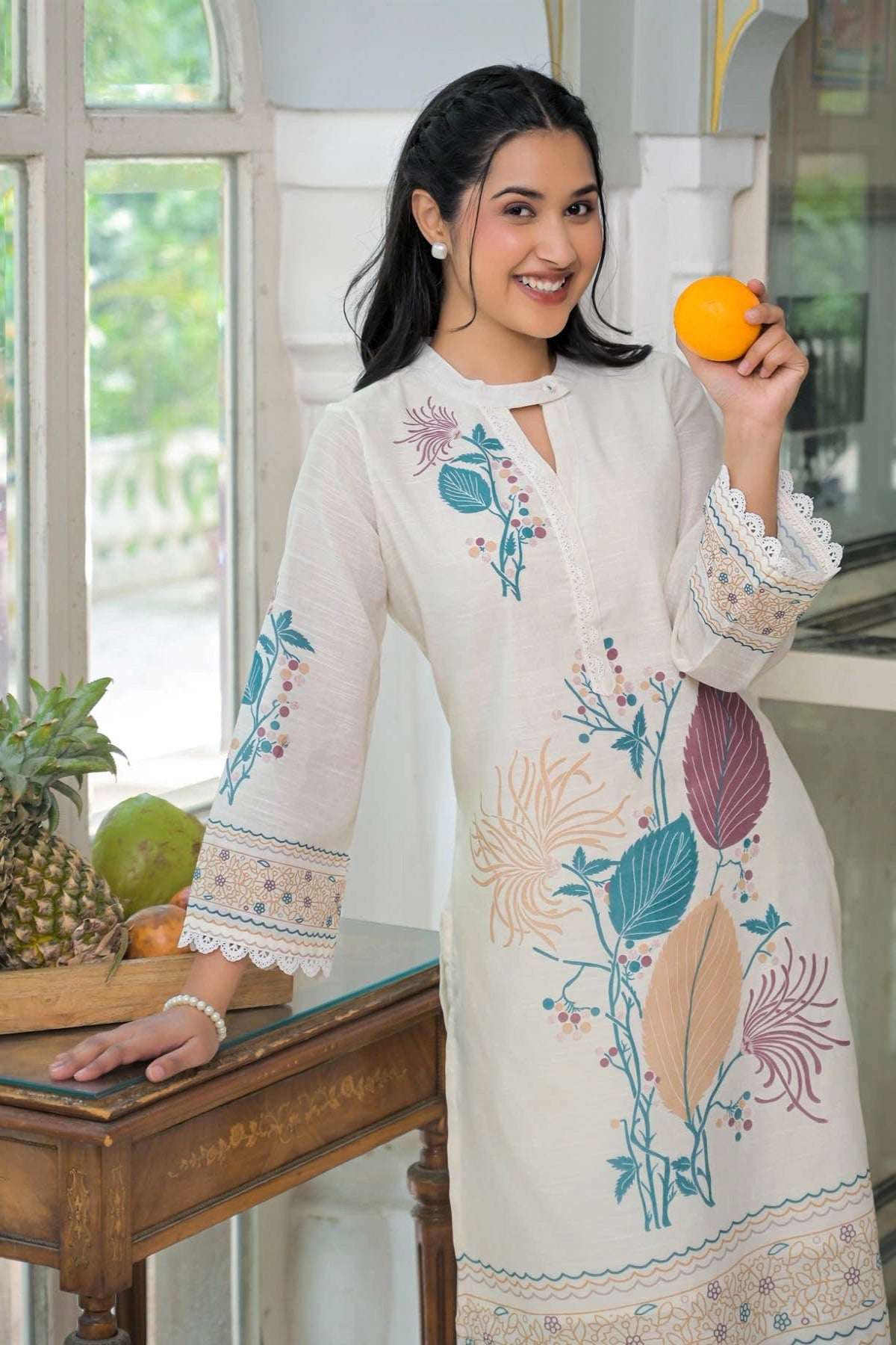 Woman in a white embroidered dress holding an orange indoors.