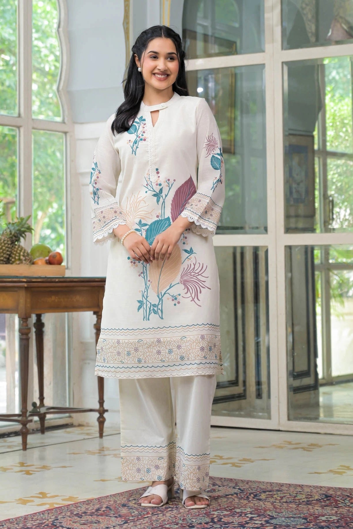 Woman wearing a white embroidered outfit in a room with large windows.