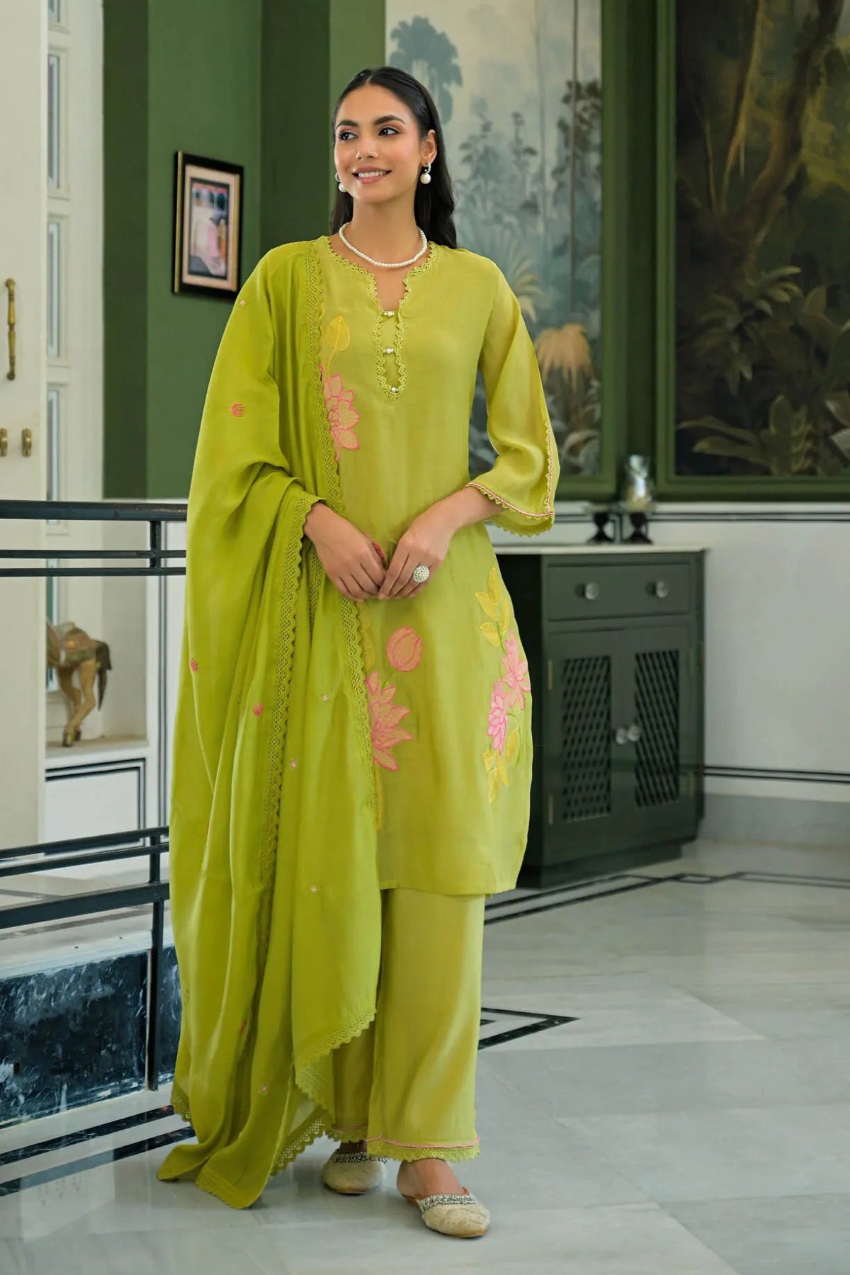 Woman in a green traditional outfit standing in an indoor setting with decorative elements.