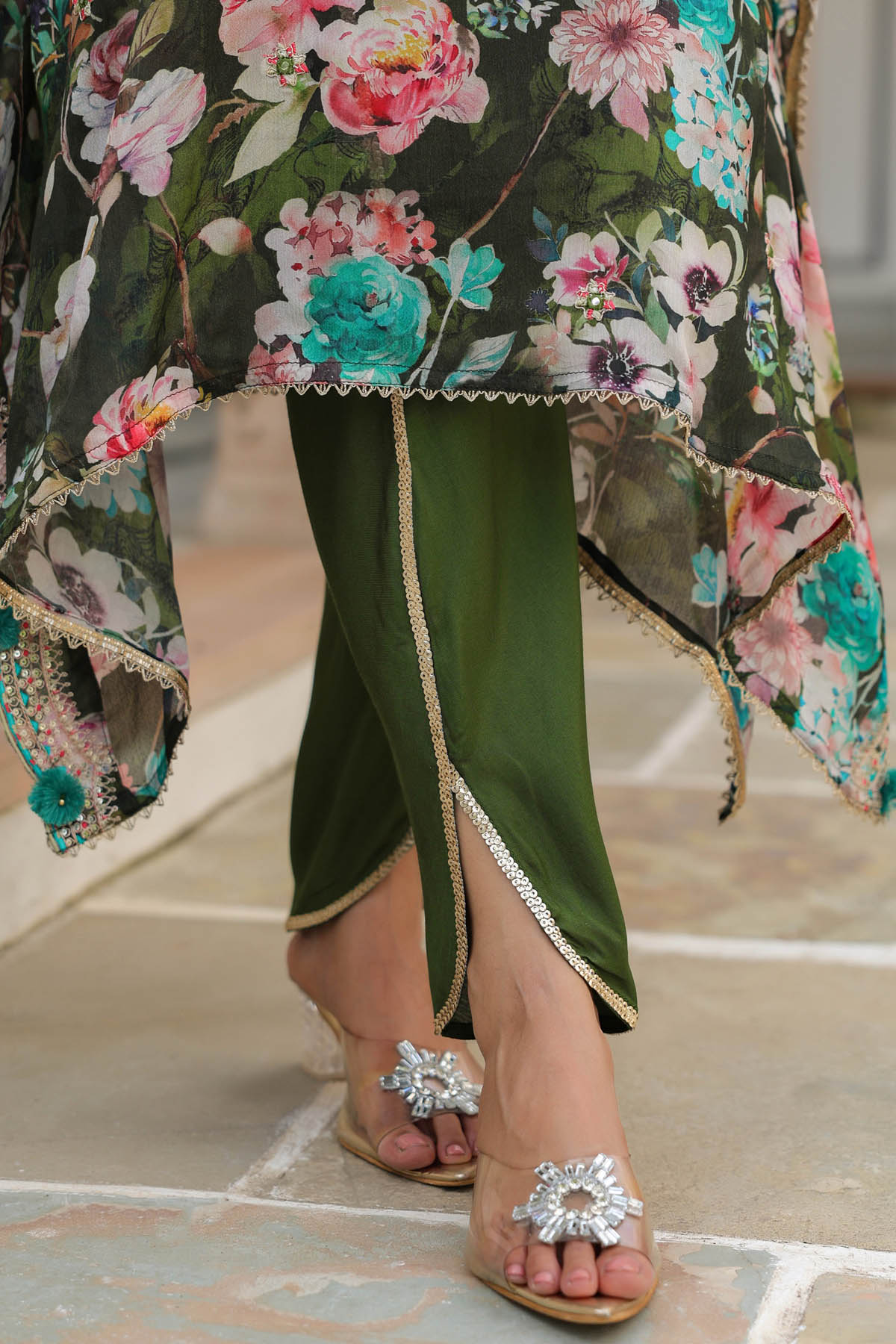 Traditional outfit with floral top and green pants, paired with embellished sandals on a stone floor.