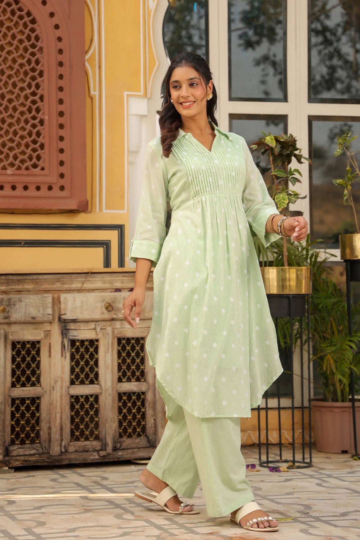 Woman in a light green traditional outfit standing outdoors with decorative elements in the background.