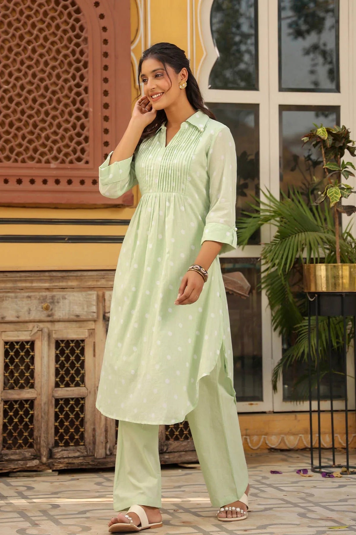 Woman in a light green traditional outfit standing outdoors with decorative elements in the background.