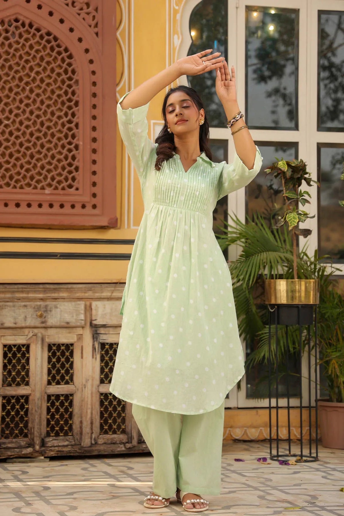 Woman in a light green traditional outfit standing in front of a decorative wall and plants.