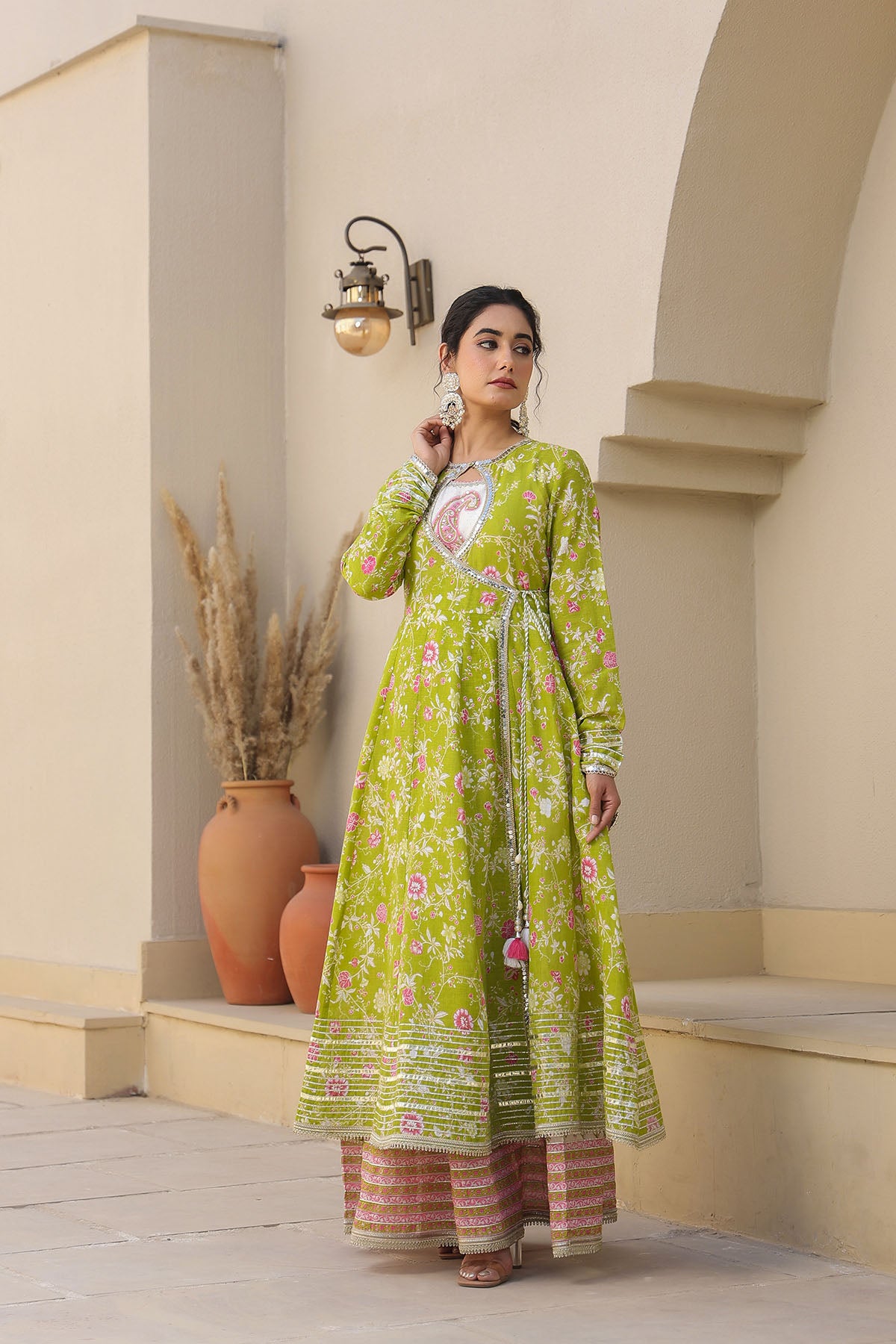 Woman wearing a green floral dress standing in an outdoor setting with beige walls and wooden steps.