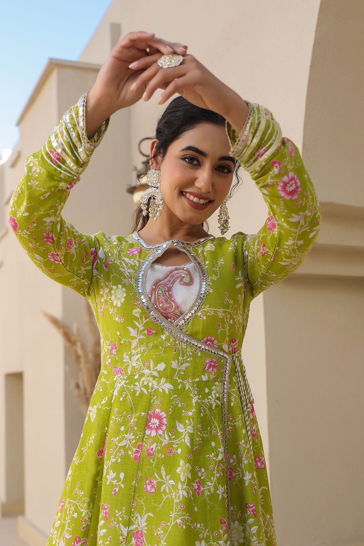 Woman in a green floral dress with jewelry, standing against a building.