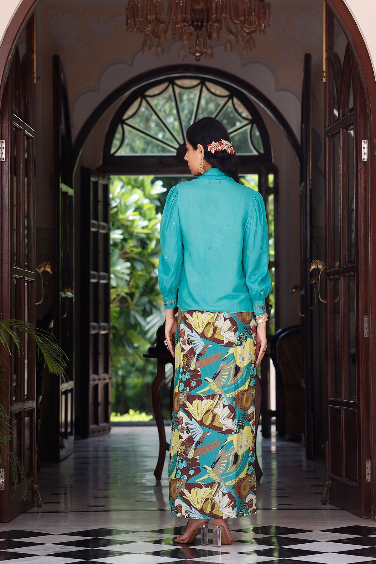 Woman in a turquoise blouse and floral skirt standing in a doorway with a decorative ceiling light.