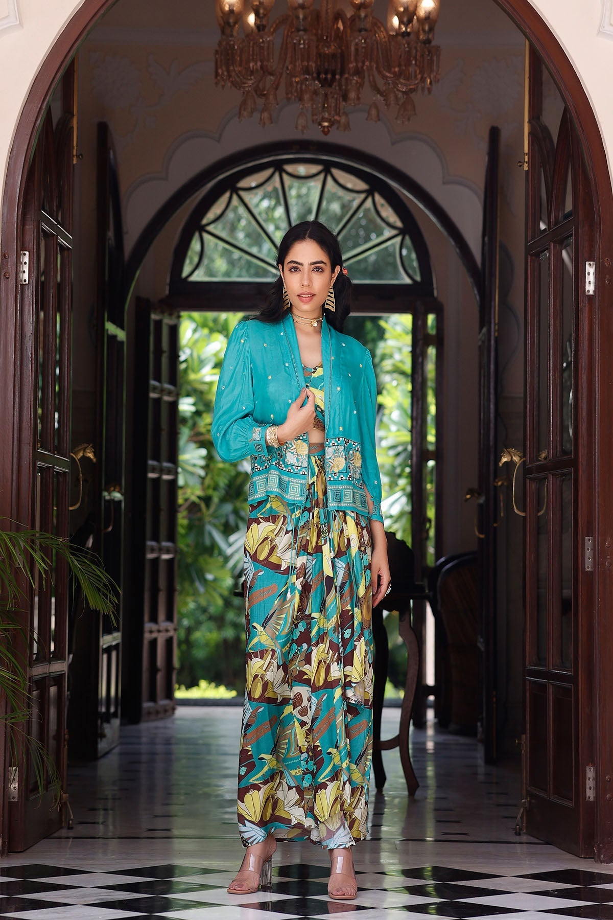 Woman in a teal jacket and colorful dress standing in a hallway with a chandelier above.