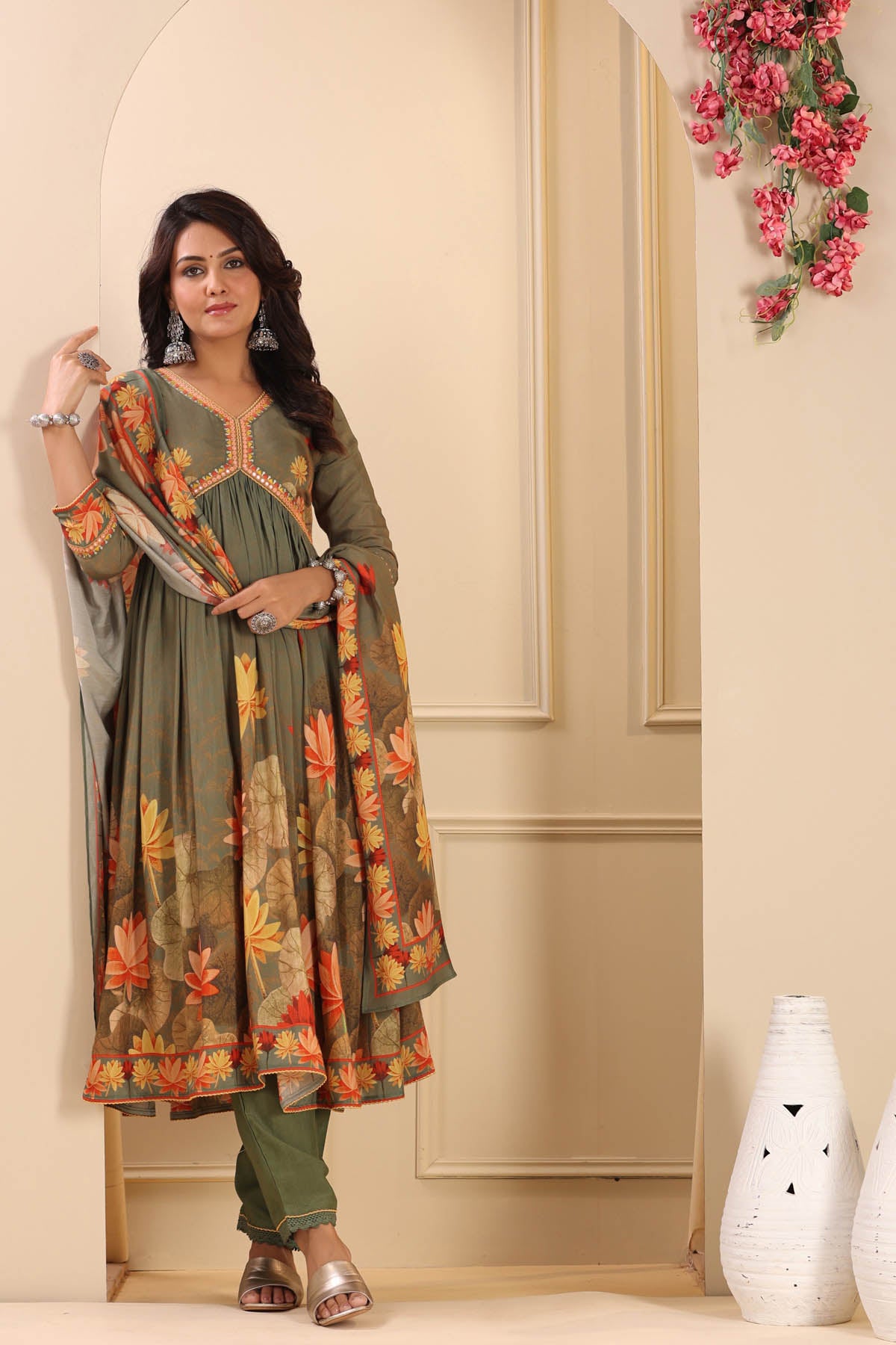 Woman wearing a green and orange traditional outfit with floral patterns in a decorated room.