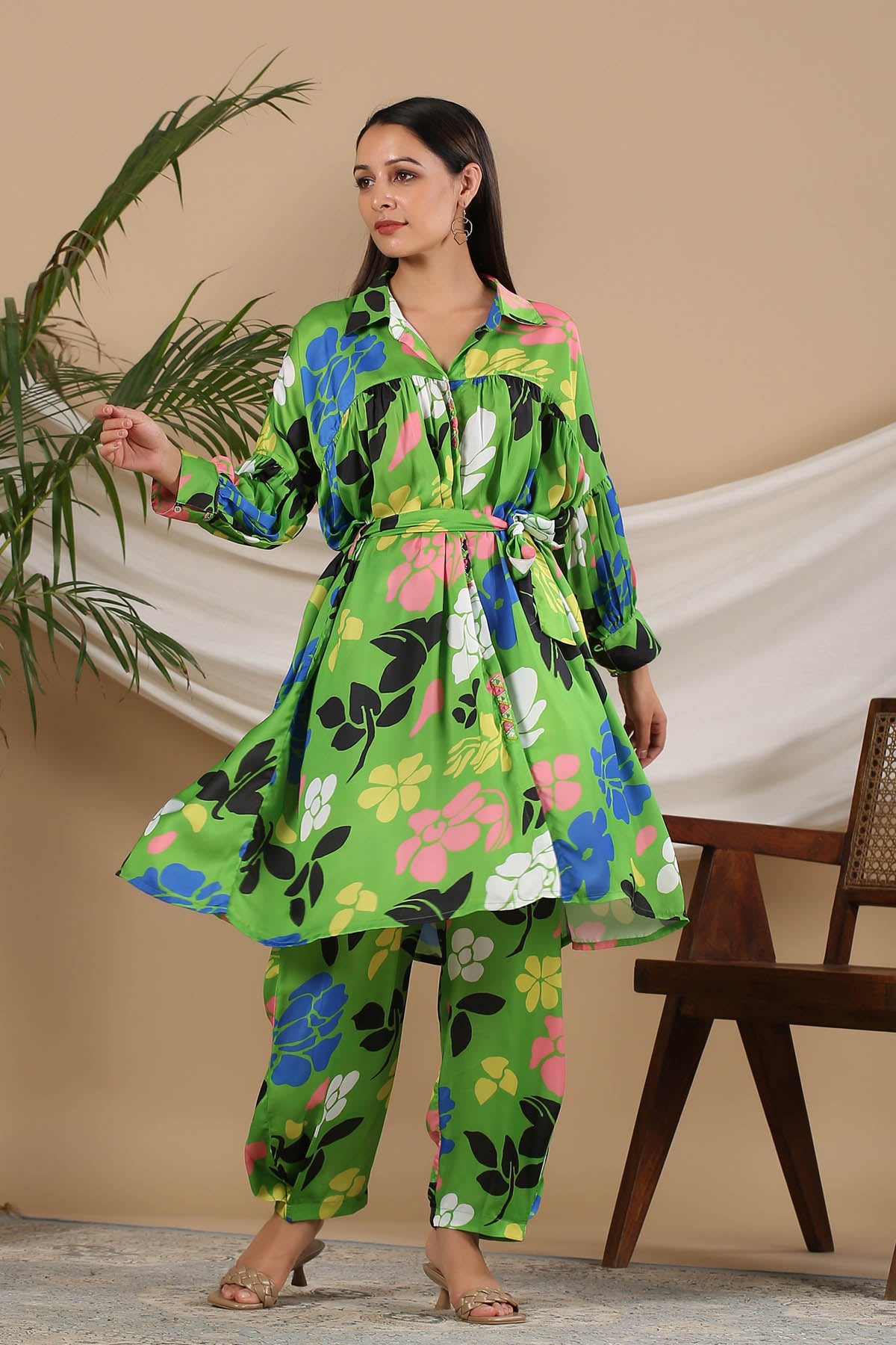 Woman wearing a green floral outfit standing indoors with a plant and chair in the background.