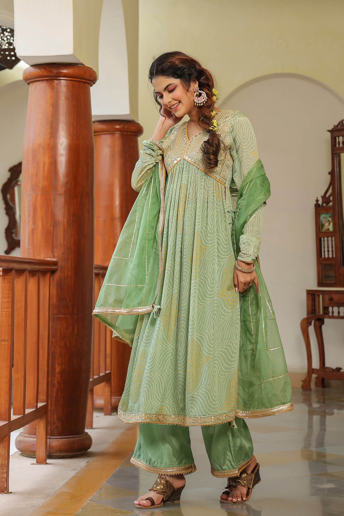 Woman in a green traditional outfit standing in a decorated room with wooden columns and furniture.