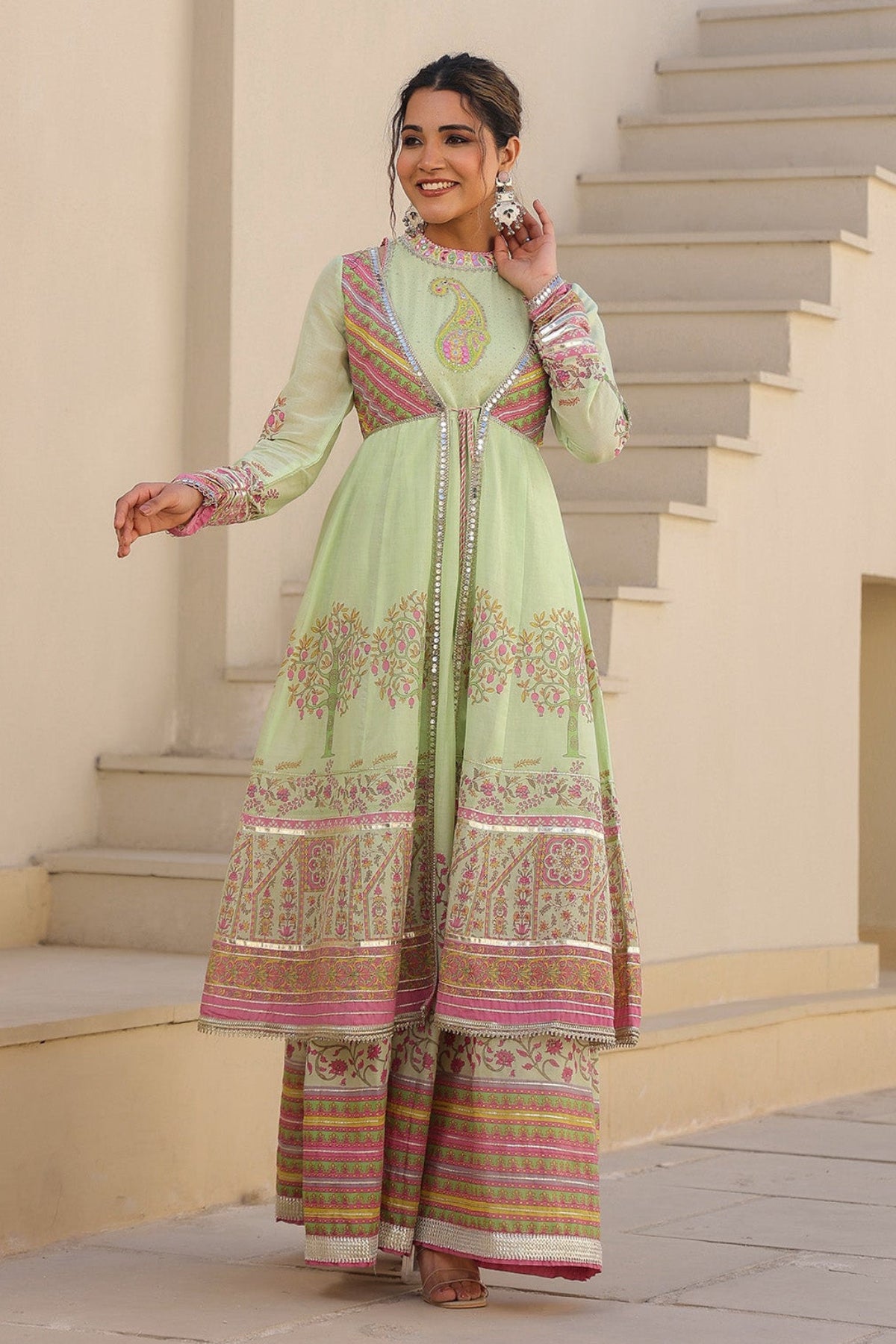 Woman in a light green traditional outfit with pink and gold patterns standing on steps.