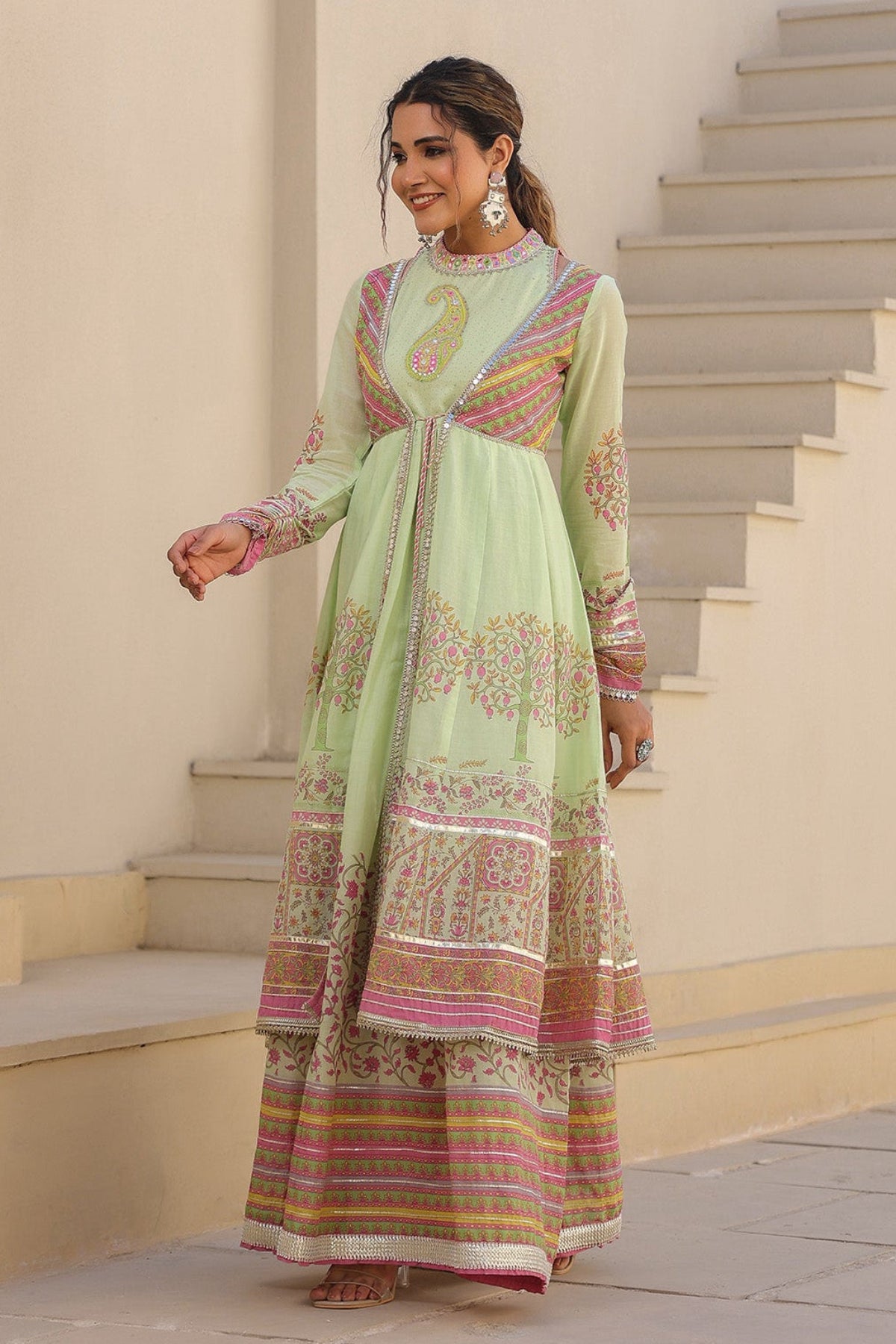 Woman in a light green traditional outfit with floral patterns standing on a staircase.