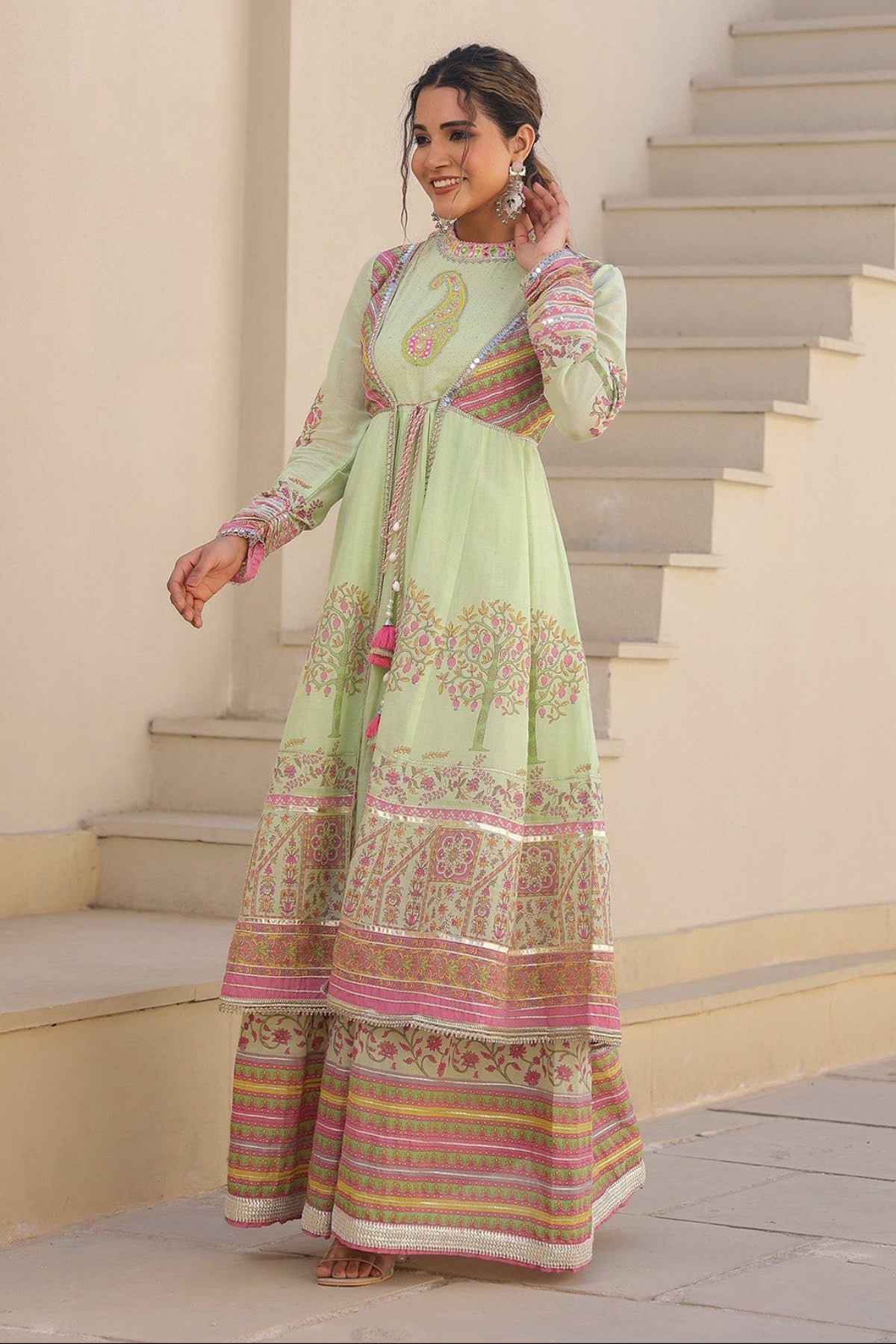 Woman in a light green traditional outfit with pink and gold patterns standing on a staircase.