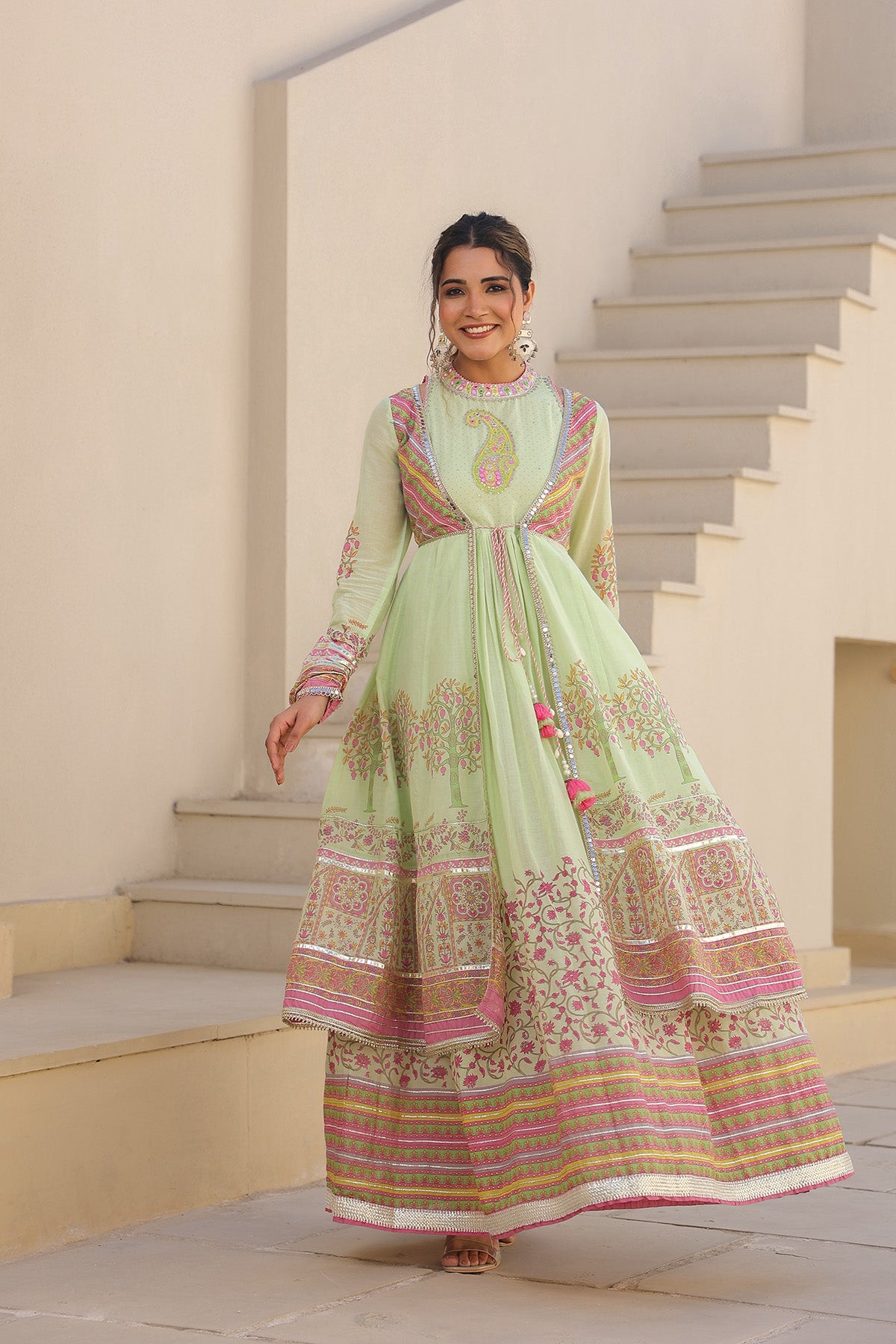Woman in a light green traditional outfit with pink and gold patterns standing on steps.