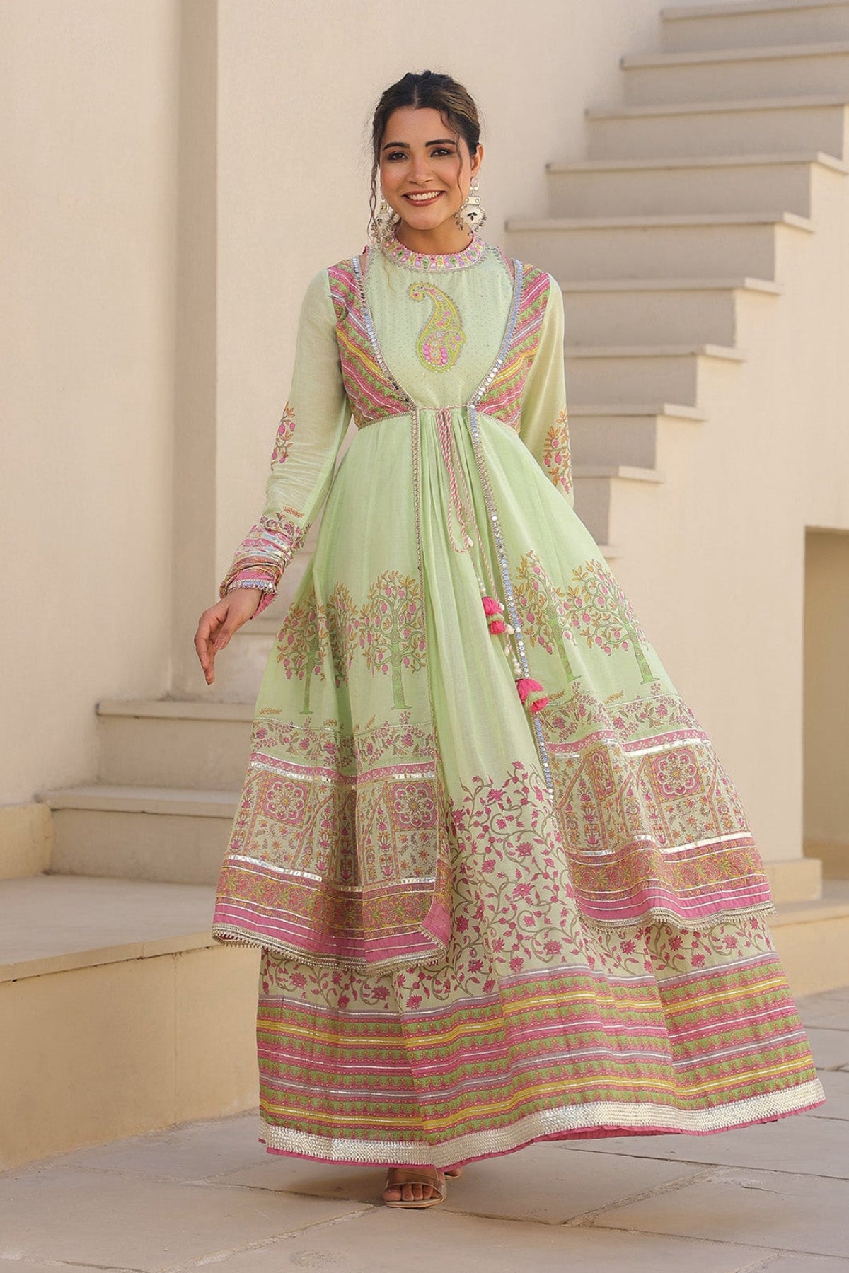 Woman in a light green traditional outfit with pink and gold patterns standing on steps.