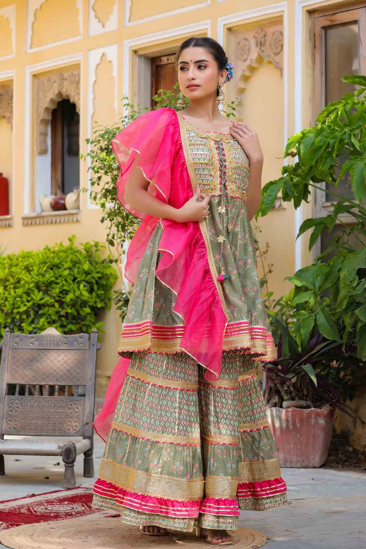 Woman in traditional green and pink outfit standing outdoors with plants and a bench in the background