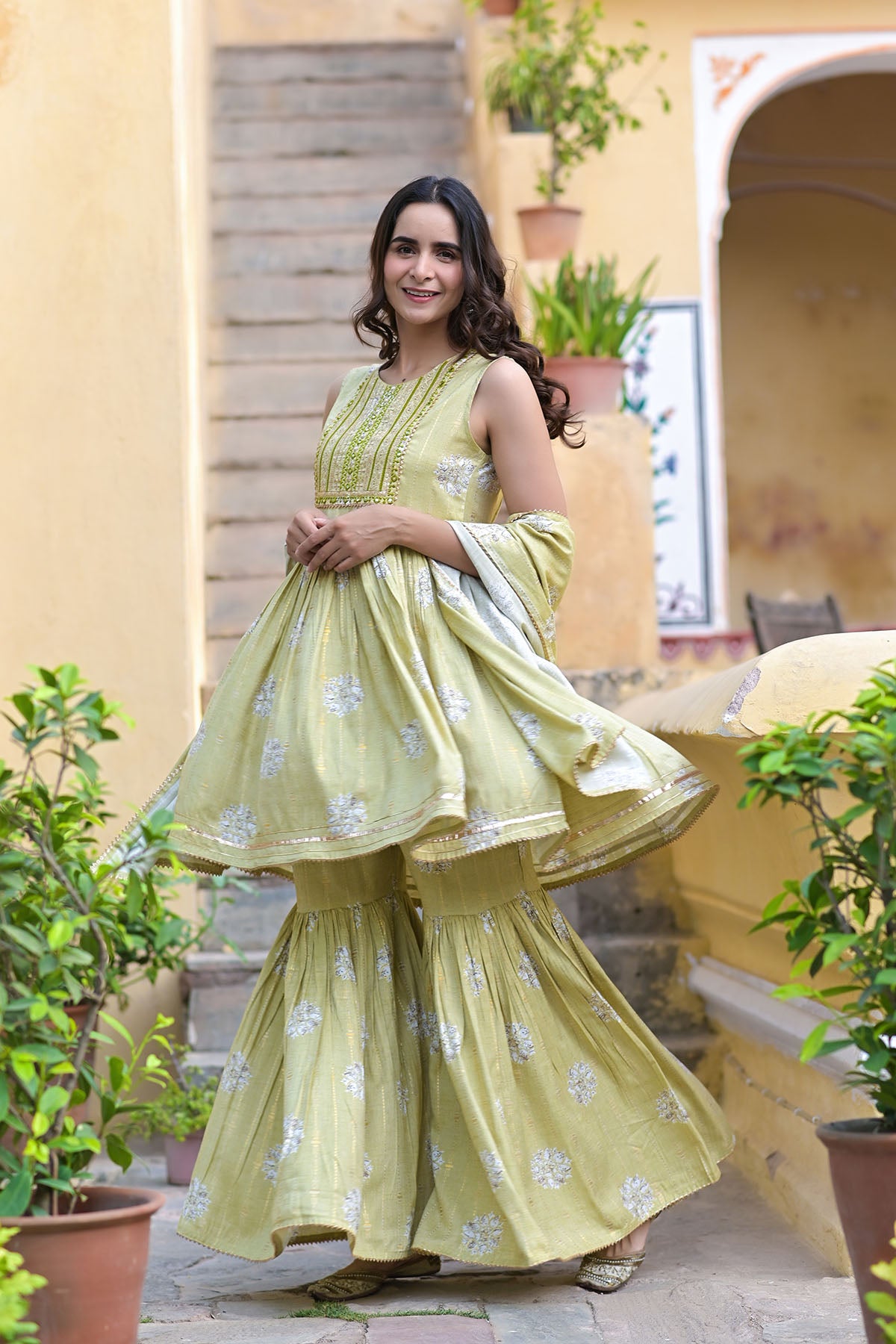 Woman in a light green traditional outfit standing outdoors with plants and a building in the background